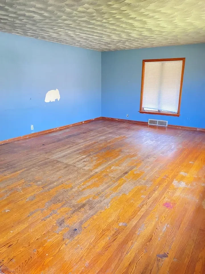 Empty room with blue walls, wooden floor with worn patches, window with closed blinds, and ceiling with textured pattern.