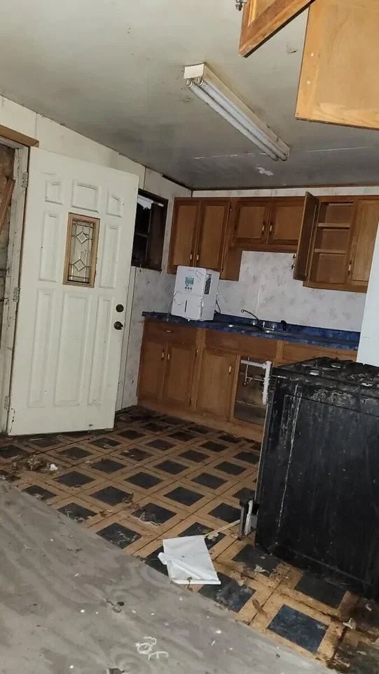 Kitchen with damaged flooring, wooden cabinets, microwave, and black stove, door with decorative glass panel, ceiling light fixture, and open cabinet above the stove.