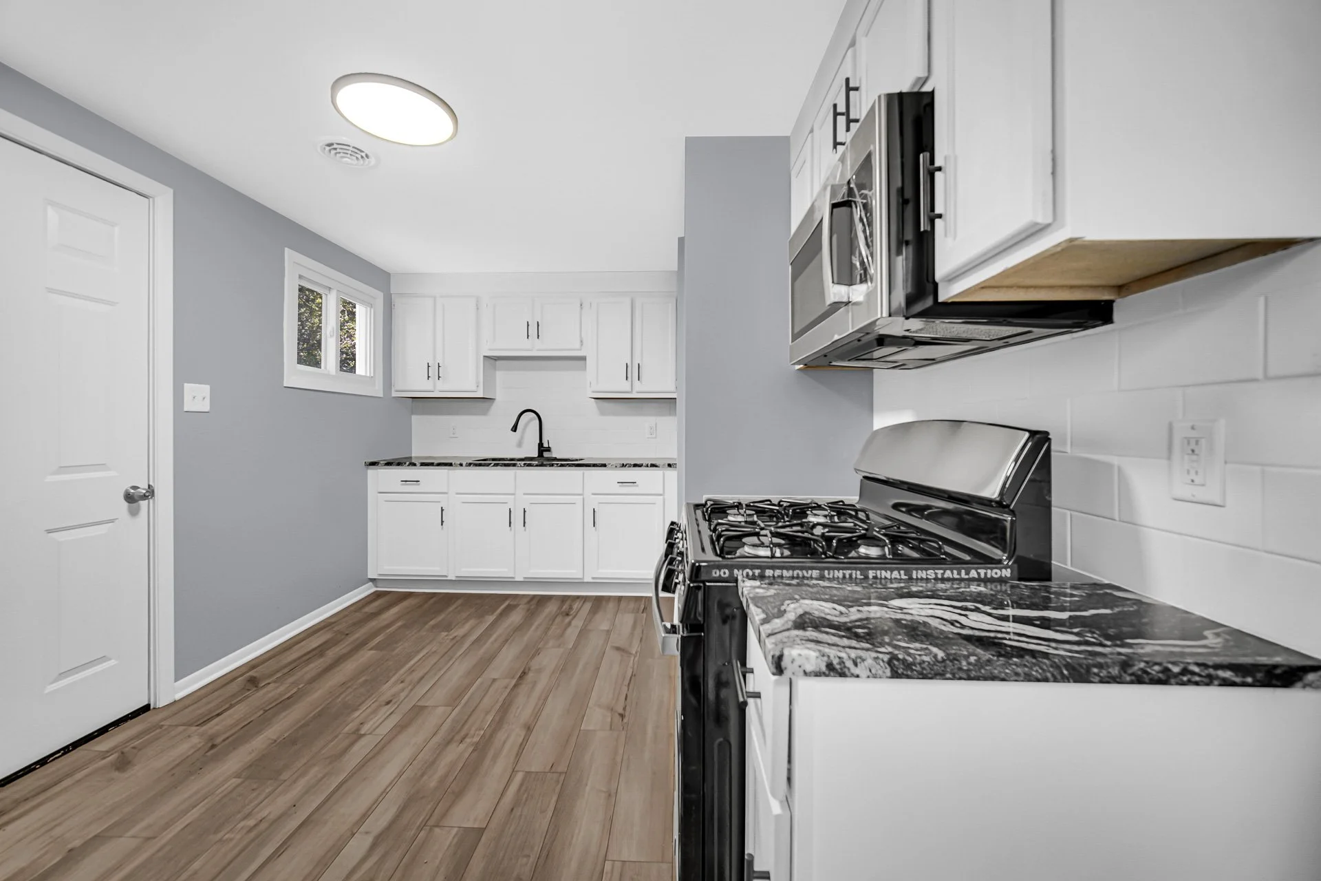 Modern kitchen with white cabinets, black hardware, gray walls, wooden laminate flooring, stainless steel stove, microwave, and black faucet at a single-basin sink.