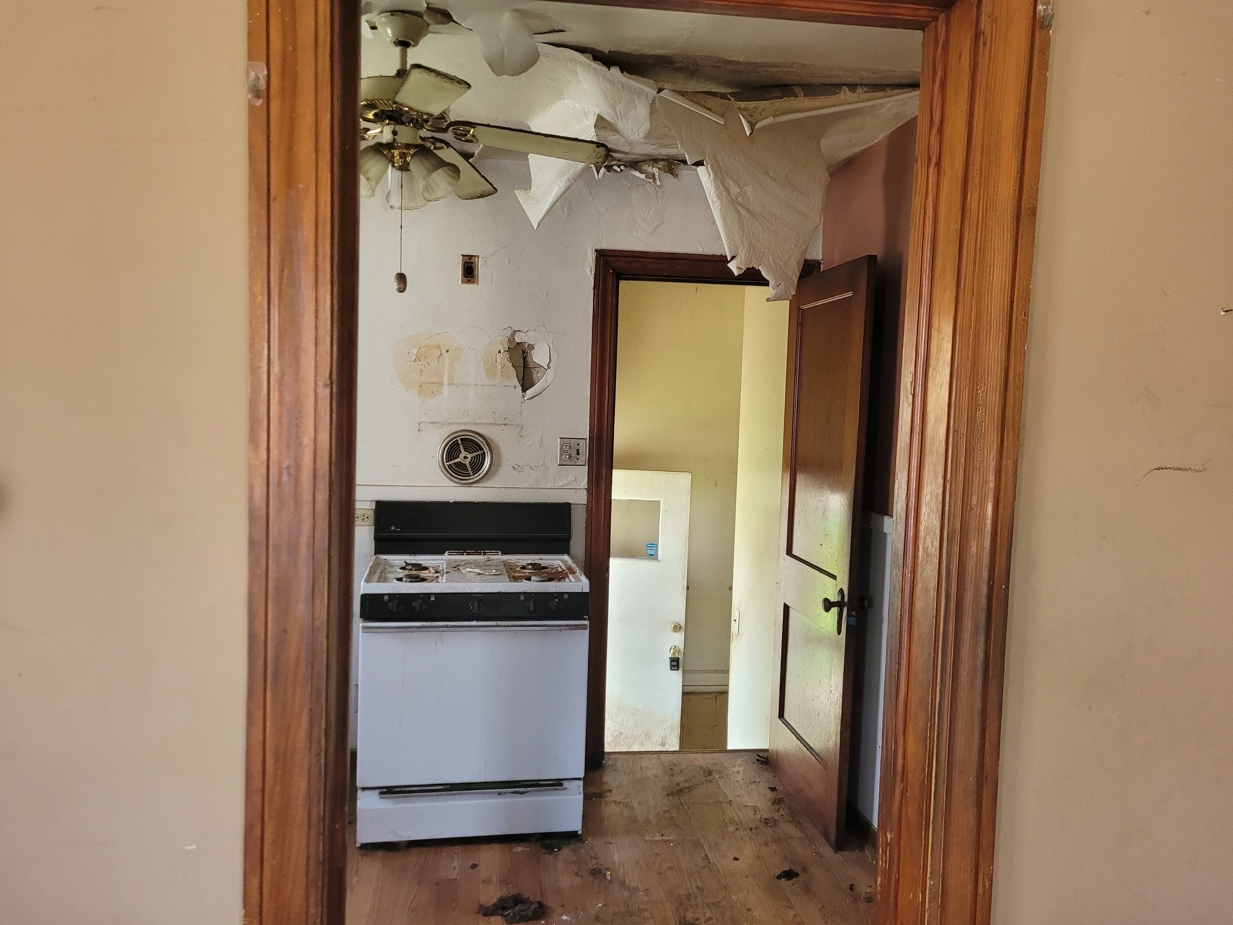 Snapshot of an old, messy kitchen with damaged walls, a ceiling fan, an air vent, and a stove. The room is viewed through a wooden-framed doorway.