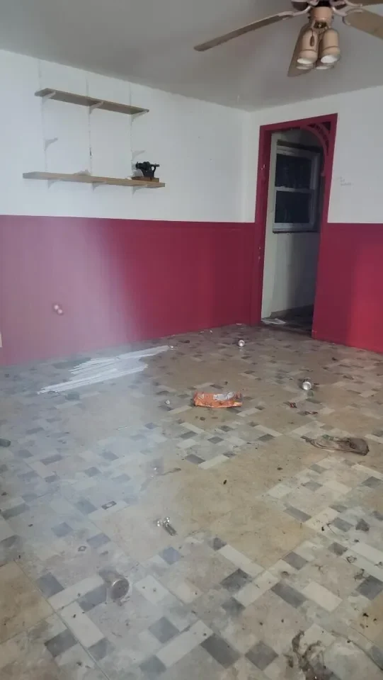 Empty room with a ceiling fan, two floating shelves on the wall, a doorway with a pink trim, and a dirty, cluttered floor with debris and a spill.