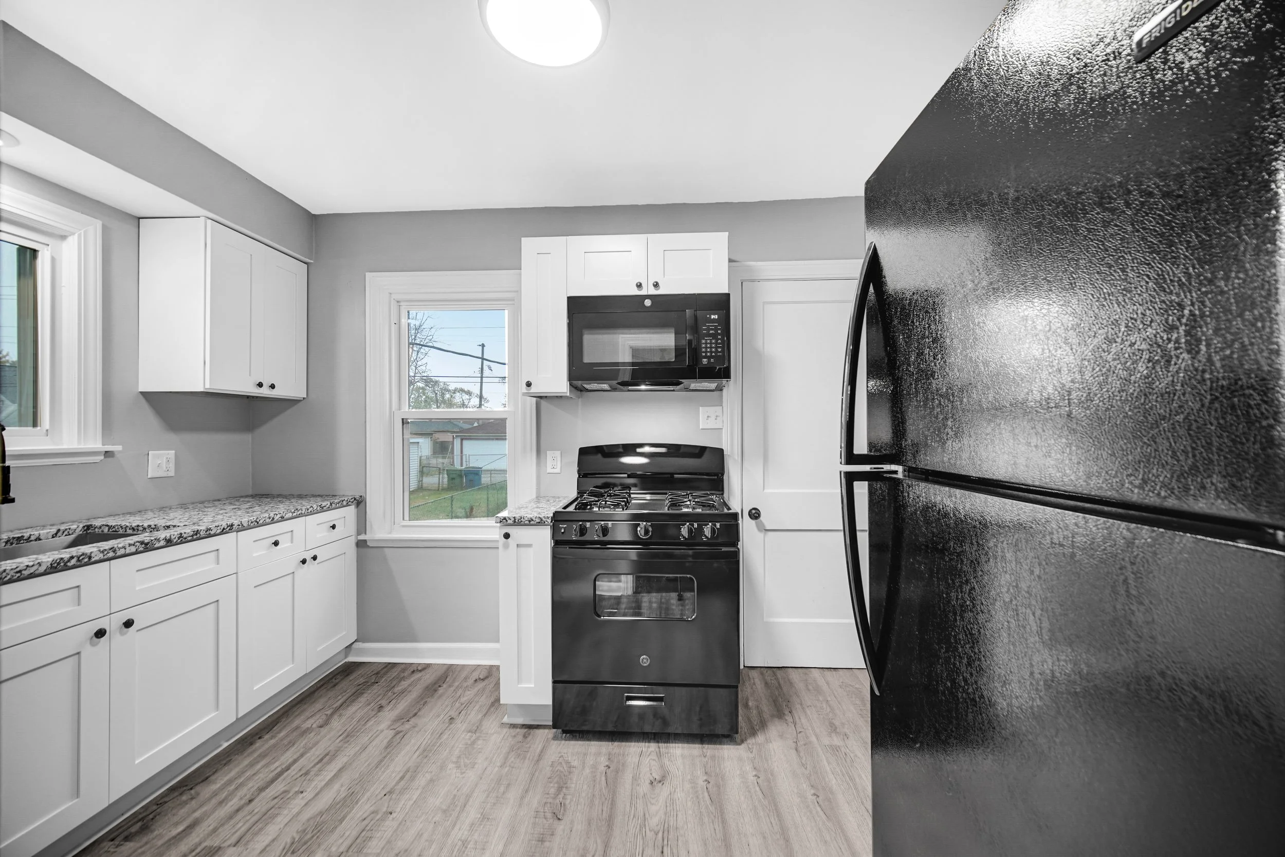 Kitchen with white cabinets, black appliances, a window showing outdoor trees and houses, and hardwood floors.