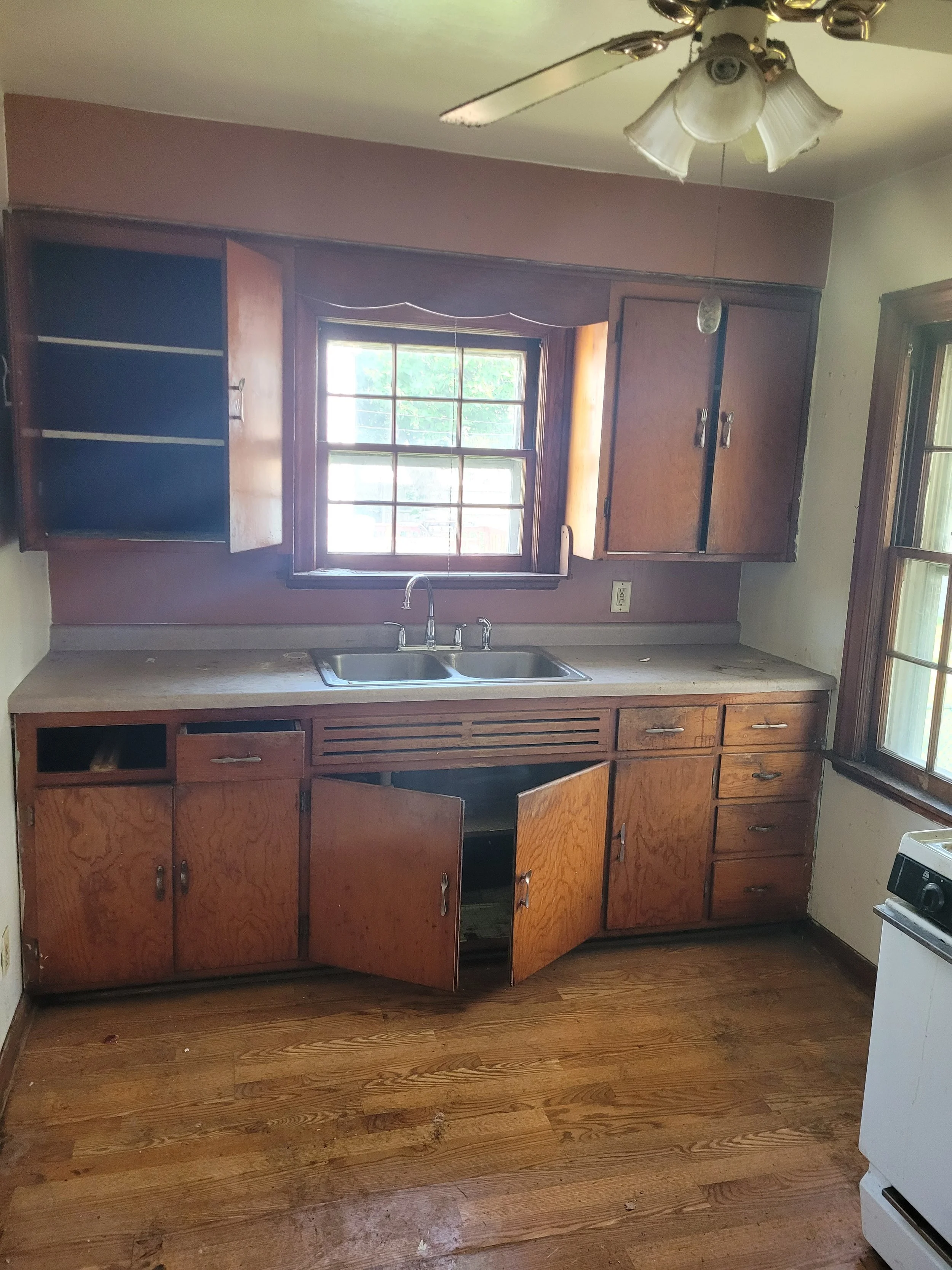 A kitchen with wooden cabinets, a window above the sink, a ceiling fan with glass light covers, and hardwood flooring.