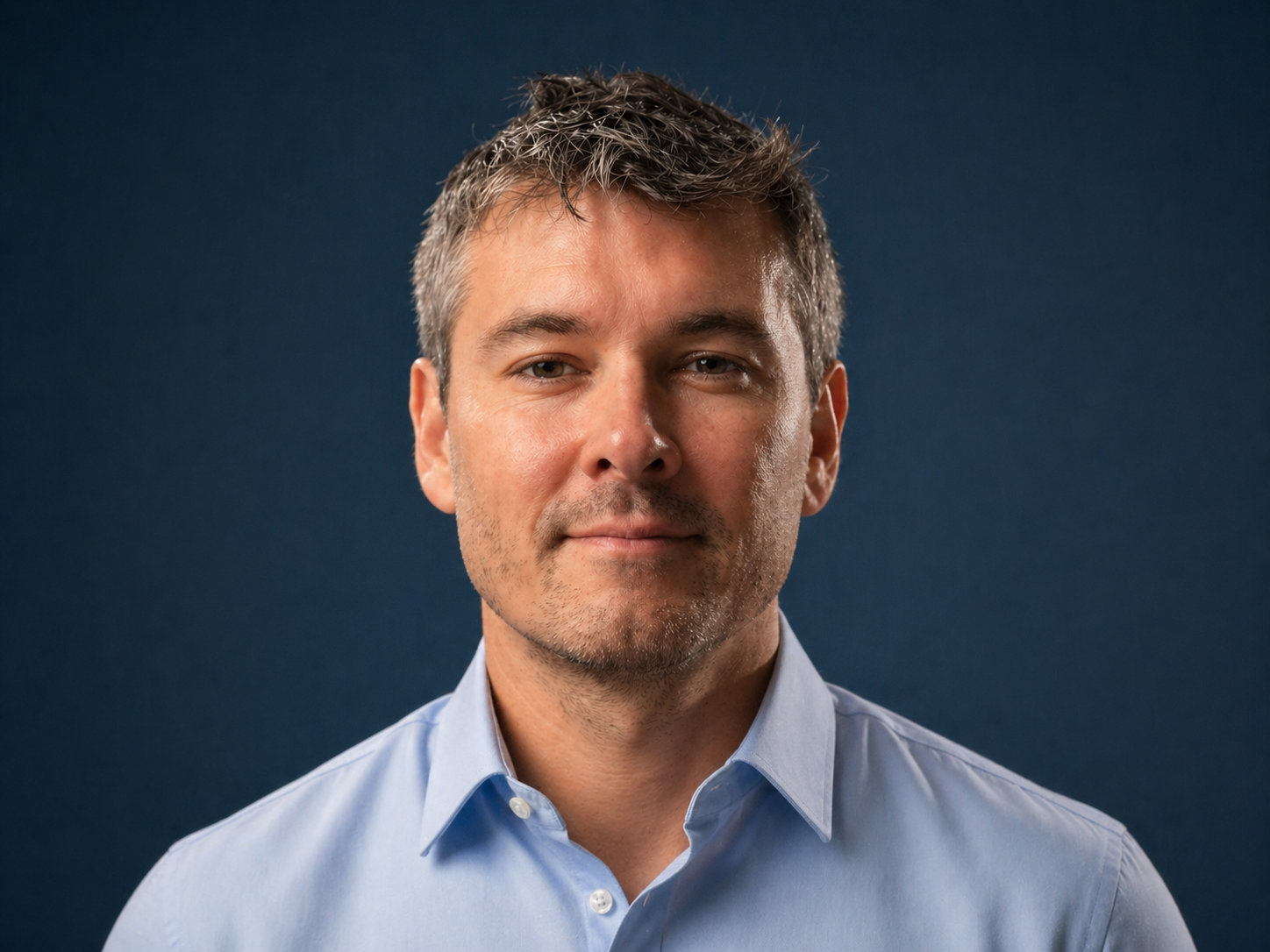 A man with short, wavy, salt-and-pepper hair wearing a light blue dress shirt against a dark blue background.