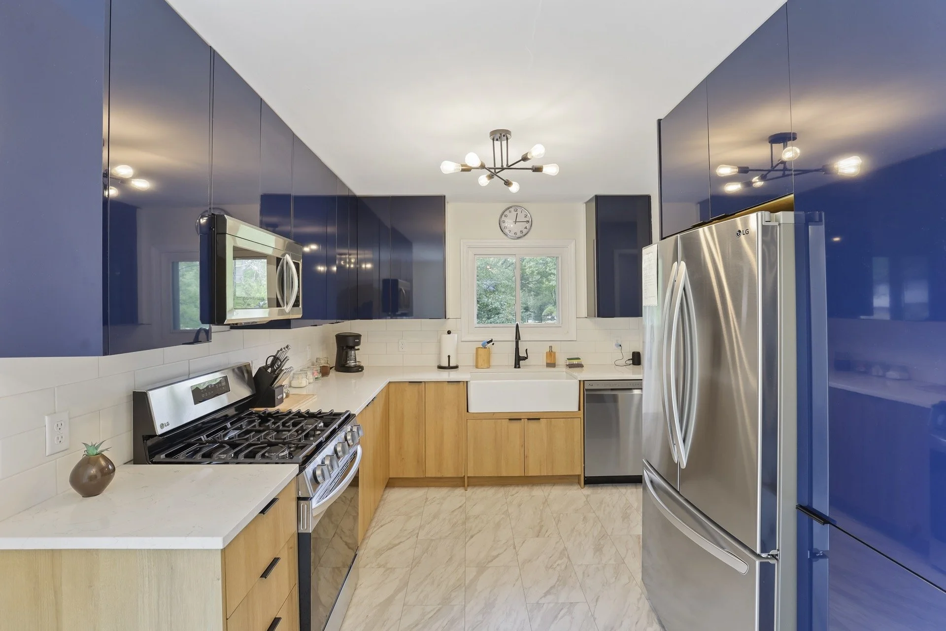 Modern kitchen with blue glossy upper cabinets, wooden lower cabinets, stainless steel appliances including a refrigerator, microwave, and stove, white tiled backsplash, white countertops, a window over the sink, a clock above the window, and decorative items like a pineapple-shaped vase and a coffee maker.