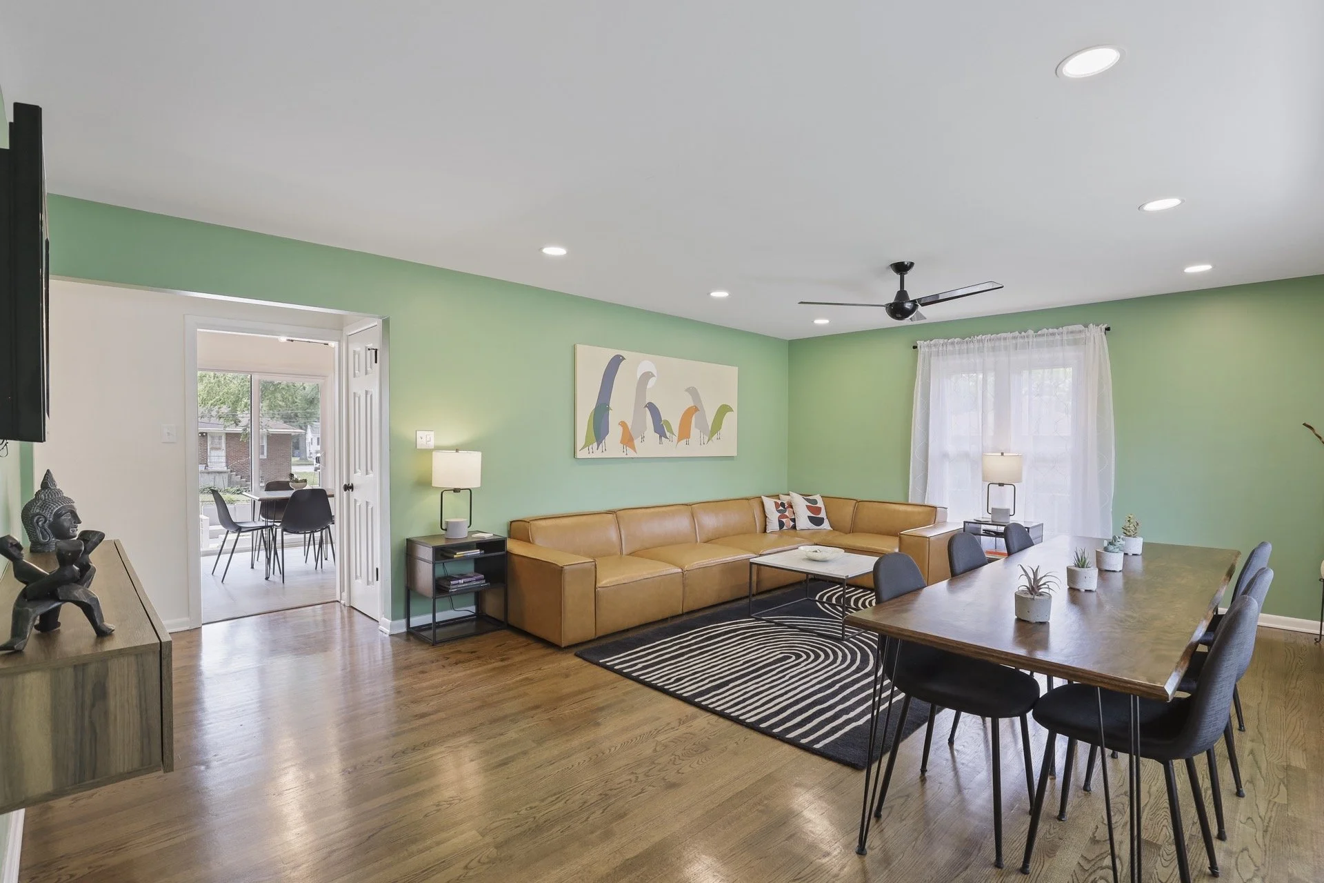 A living room with a green accent wall, a tan leather sofa, a black and white striped rug, a dining table with potted plants, and natural light coming through a window with white curtains.