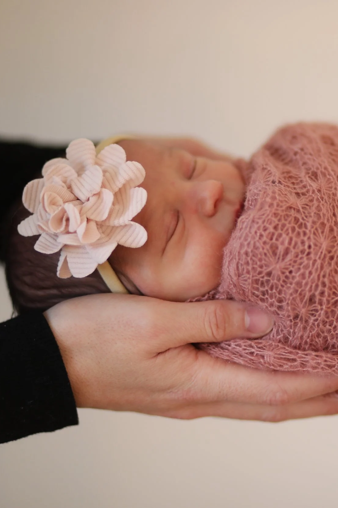 Newborn baby sleeping peacefully while being held gently by an adult's hands, wrapped in a pink knitted blanket, with a pink scrunchie on its head.
