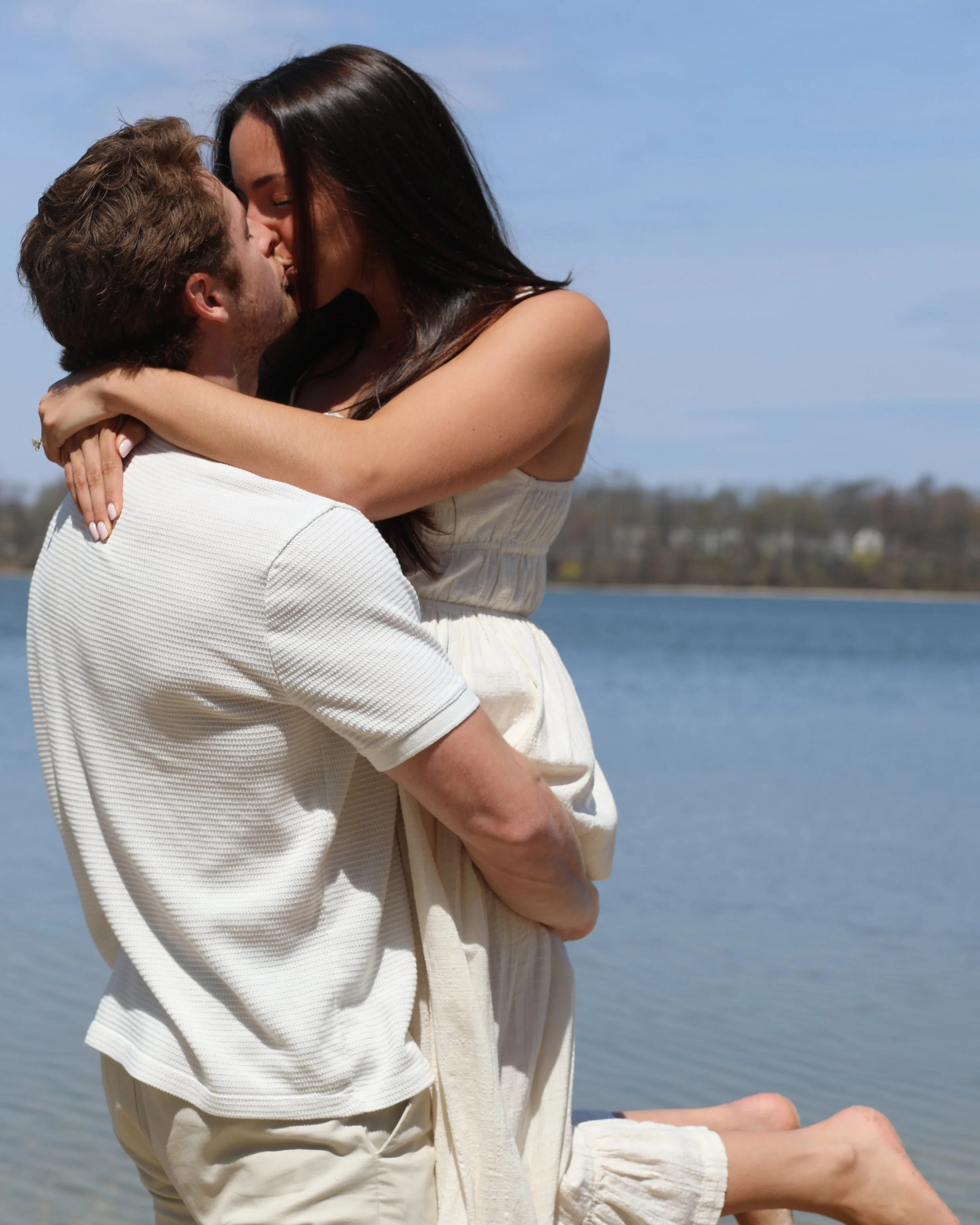 A couple near a body of water, embracing and kissing with a clear sky and distant shoreline in the background.