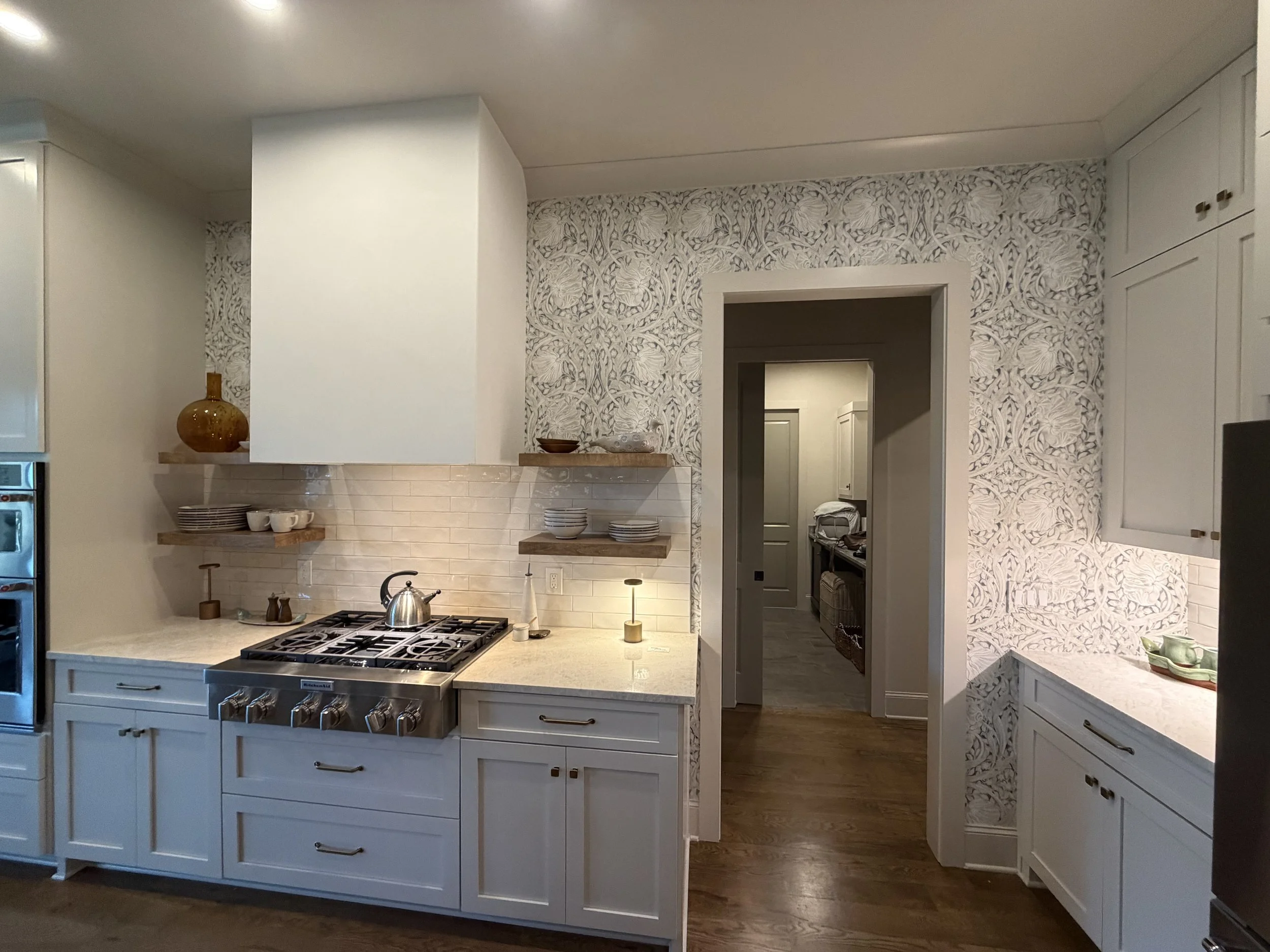 A modern kitchen with white cabinets, a gas stove, open shelves with dishes, and a patterned wall with intricate floral designs.