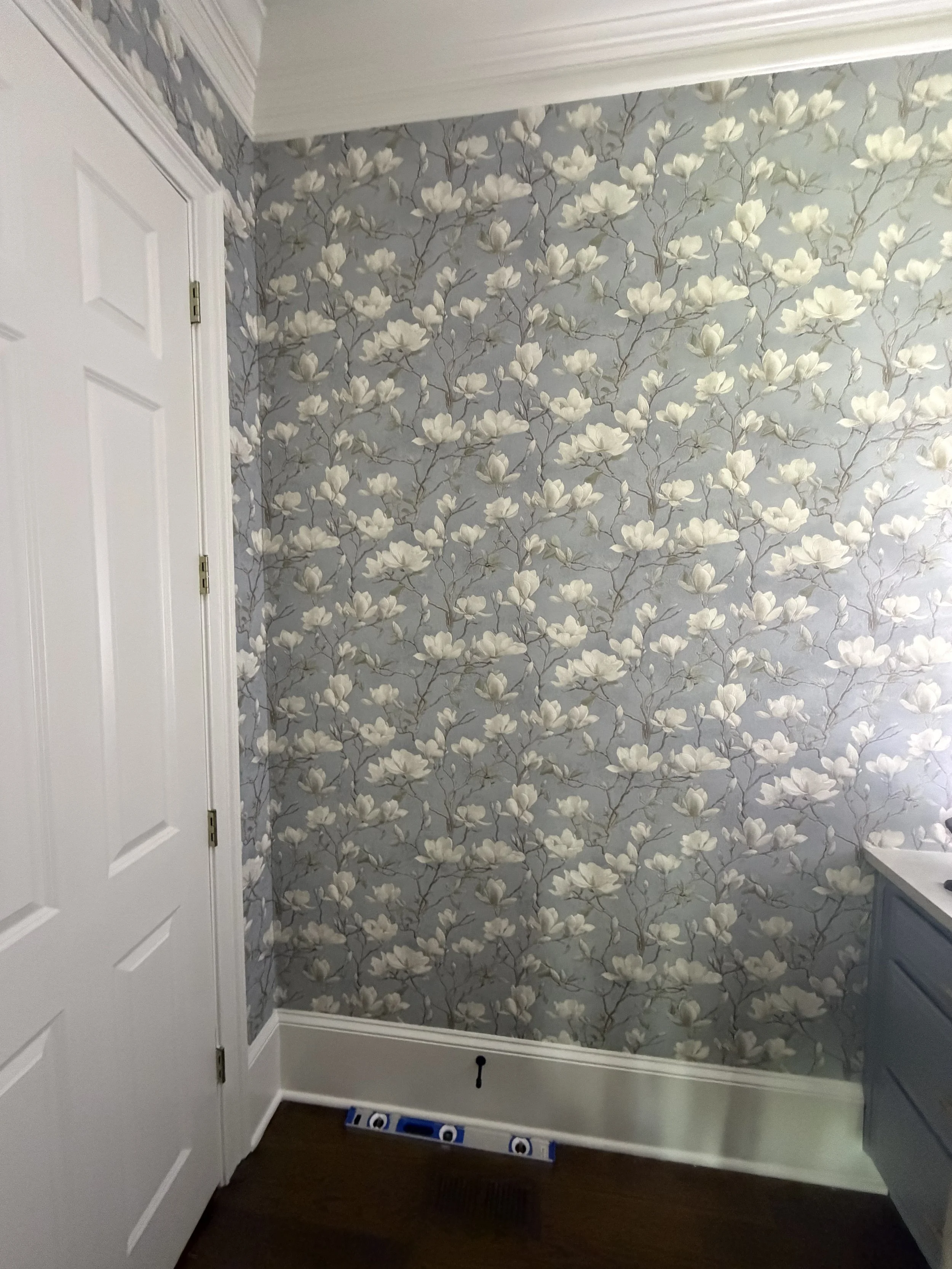 Interior corner of a room with a closed white door on the left, floral wallpaper with white flowers on a grey background on the walls, a white baseboard, a brown wooden floor, a blue and white box on the floor, and part of a dark grey dresser on the 