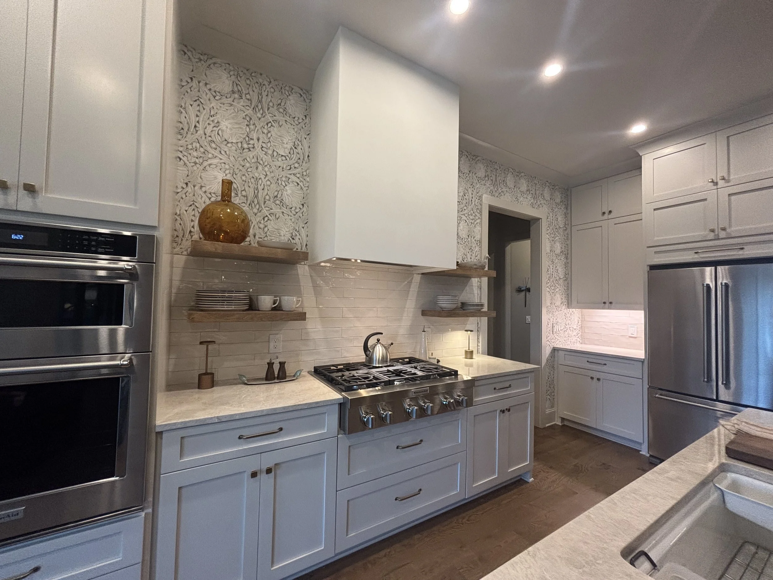 A modern kitchen with white cabinets, a stainless steel oven, and a gas stove. There are open shelves with dishes and decorative items, and the backsplash is made of beige subway tiles. The kitchen has wood flooring and recessed ceiling lights.