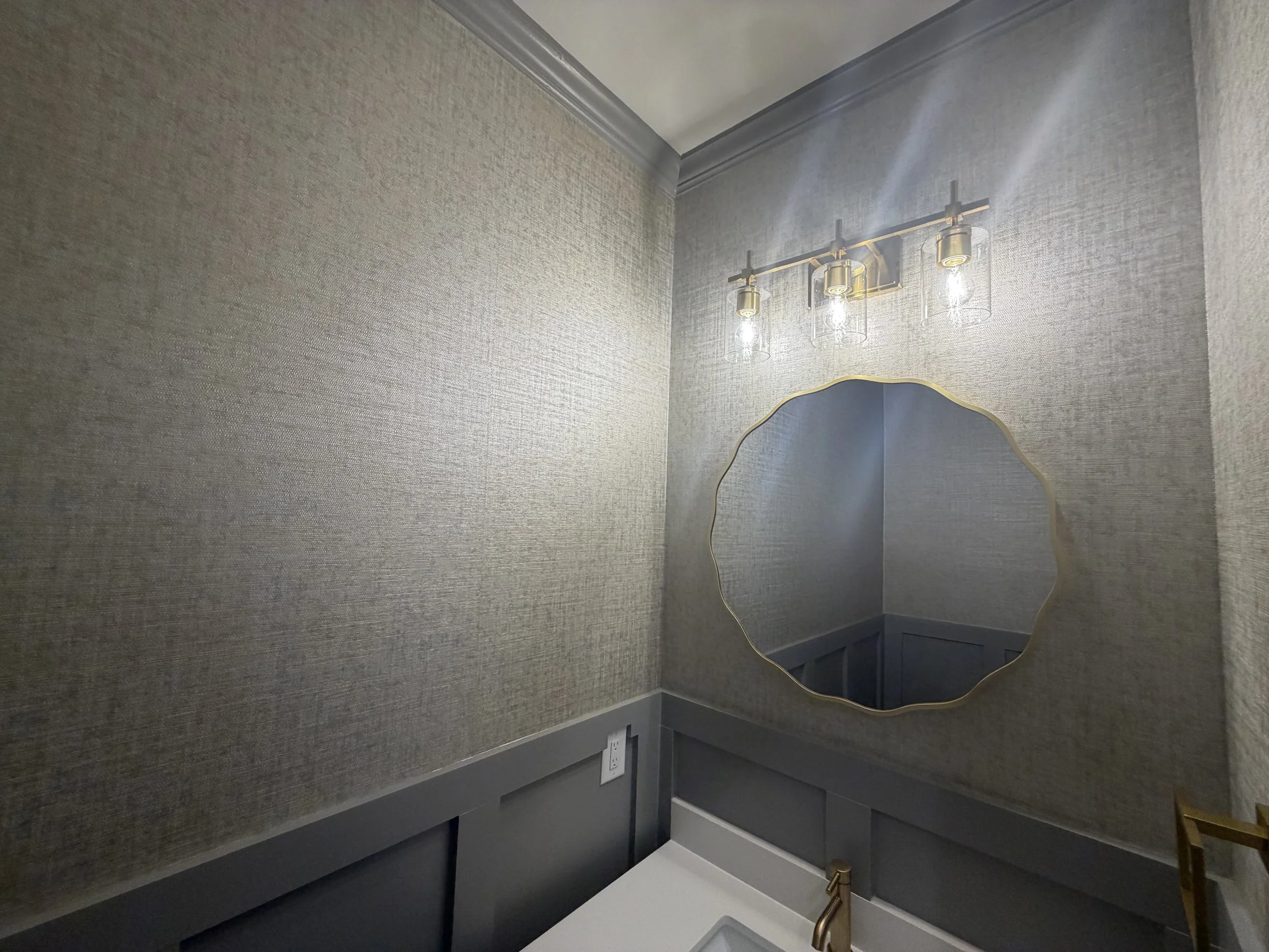 A room with gray textured walls, a gold-framed mirror, brass light fixtures with clear glass shades, and a dark gray shelf with a white surface below. There are electrical outlets on the wall near the shelf.