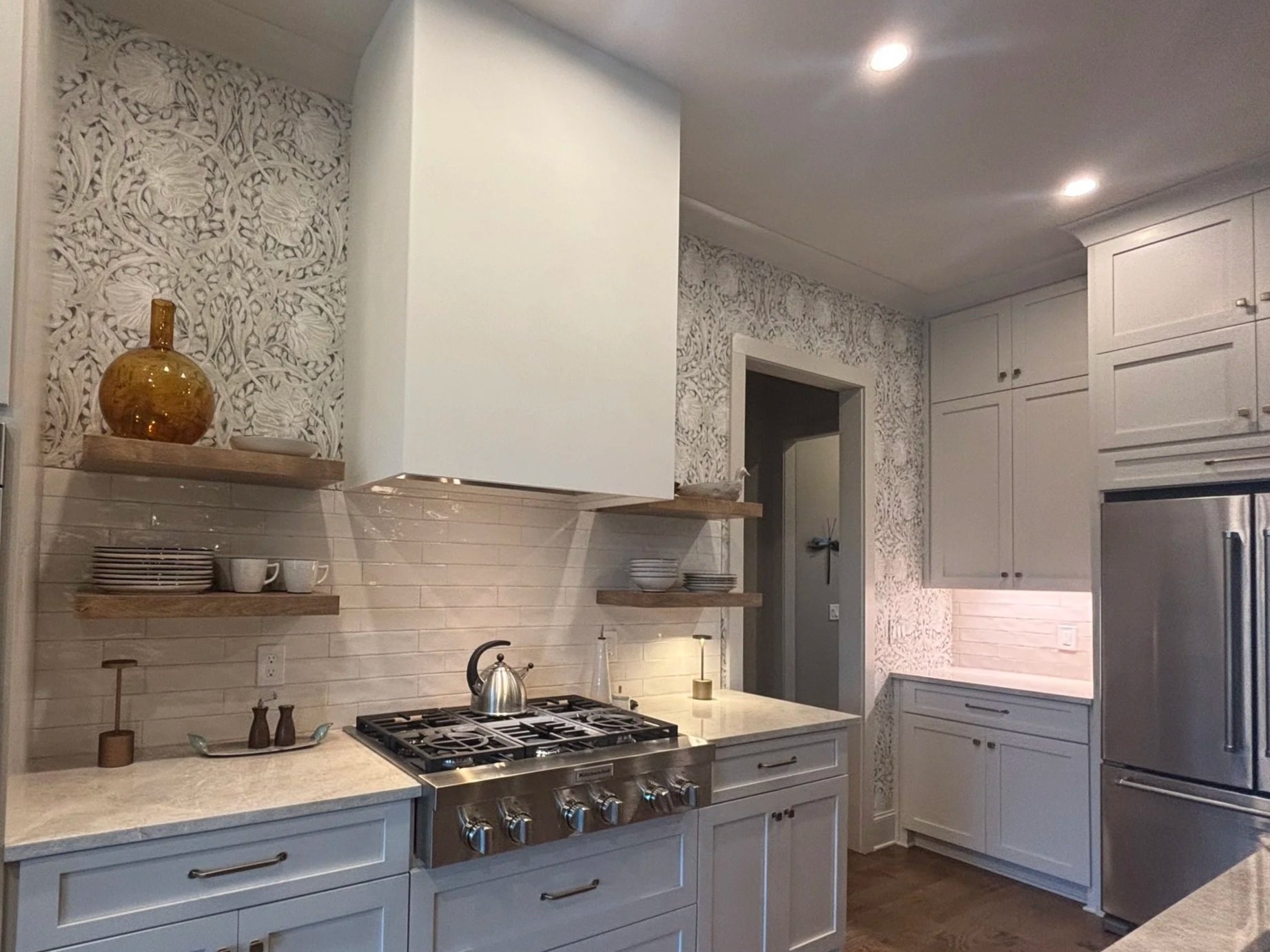 A modern kitchen with white cabinets, open shelves with dishes, a beige backsplash, and a stove with a kettle. Decorative items include a large amber vase and bowls on the shelves, with patterned wallpaper on the wall and recessed ceiling lights.