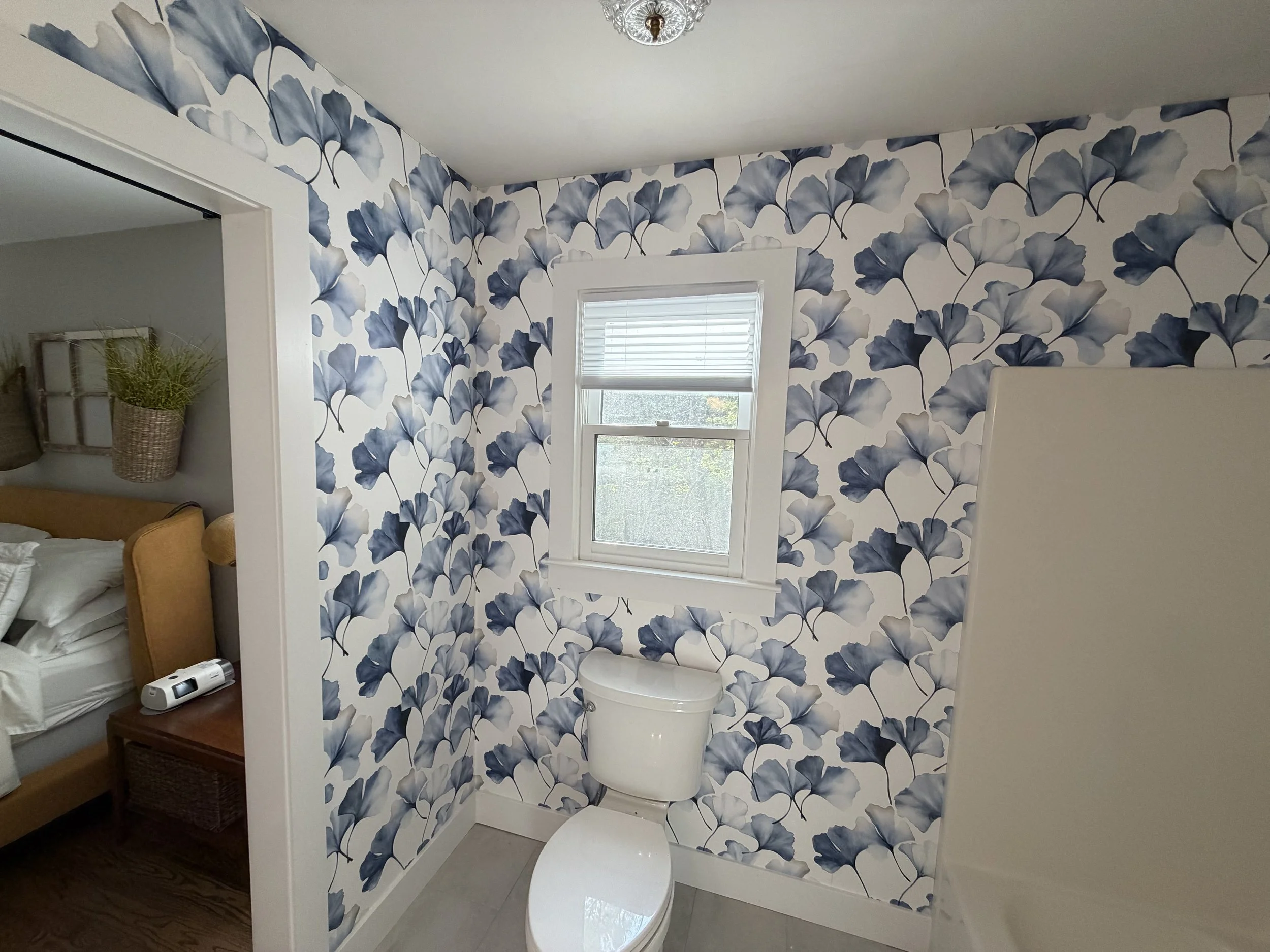 A small bathroom with floral wallpaper in shades of blue and white, a window with blinds, a white toilet, and a vase with green plants in an adjacent room.