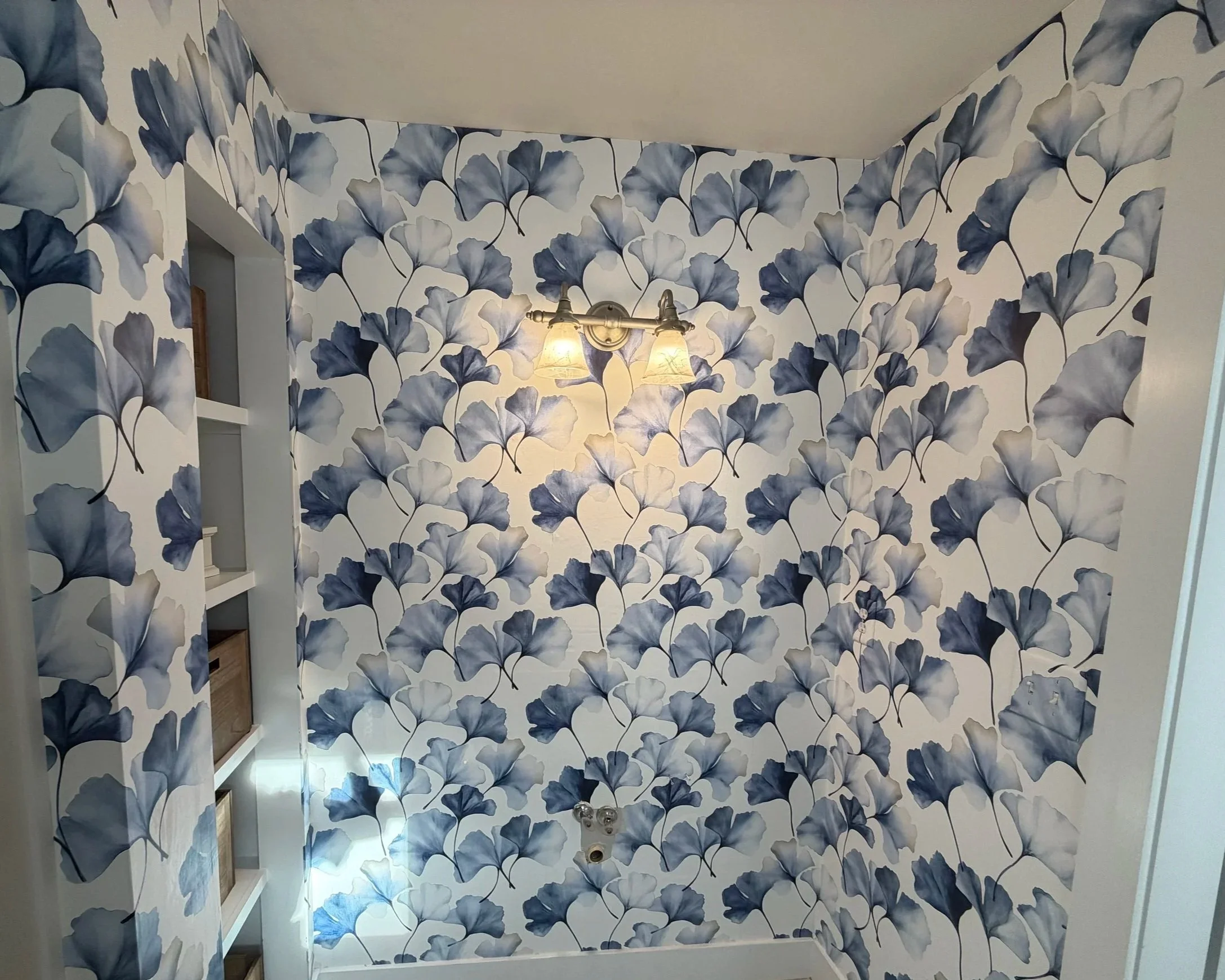 A room with wallpaper featuring a large blue and gray ginkgo leaf pattern. A wall-mounted light fixture with two lamps is turned on, casting light onto the wallpaper. A window or opening is visible on the left side. The ceiling is beige, and there is a white shelf on the left side of the image.