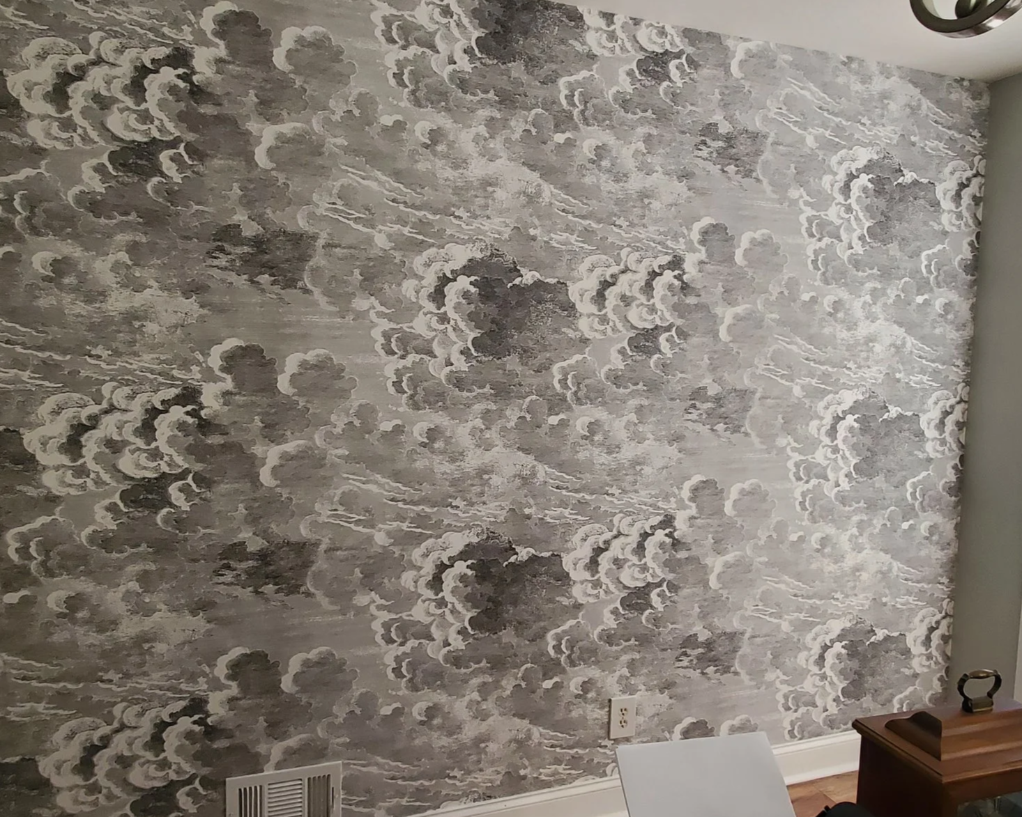 A textured wall with a stormy cloud design in shades of gray and white, with a vent and electrical outlet at the bottom and a small table with items on the right side.