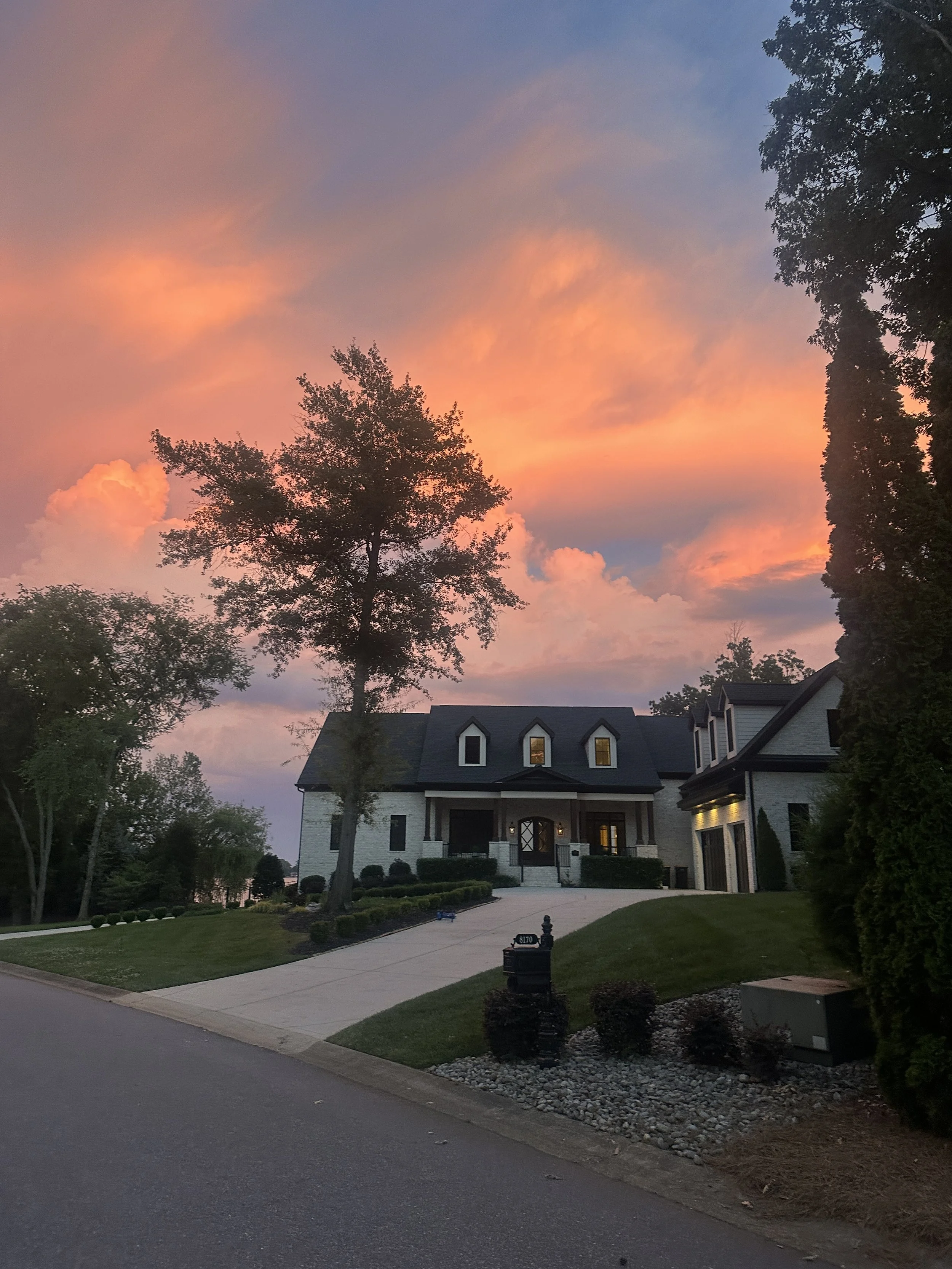 Large modern house with a white exterior, dark roof, and illuminated front porch, surrounded by trees and a well-maintained lawn, during sunset.