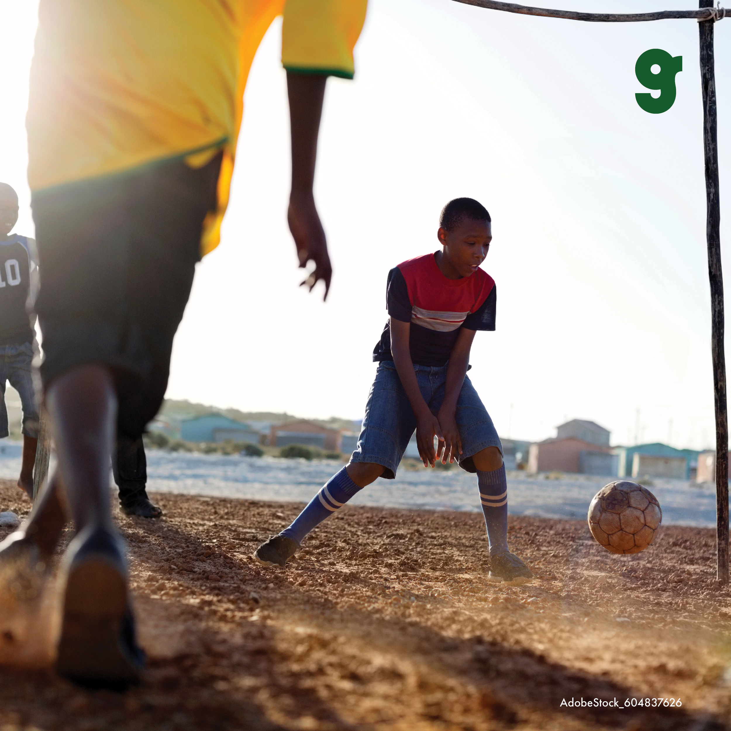 Children playing soccer on a dirt field with a goalpost, viewed from the ground level with the camera close to the ground. The sun is low, creating a backlit scene with silhouettes and a bright sky.
