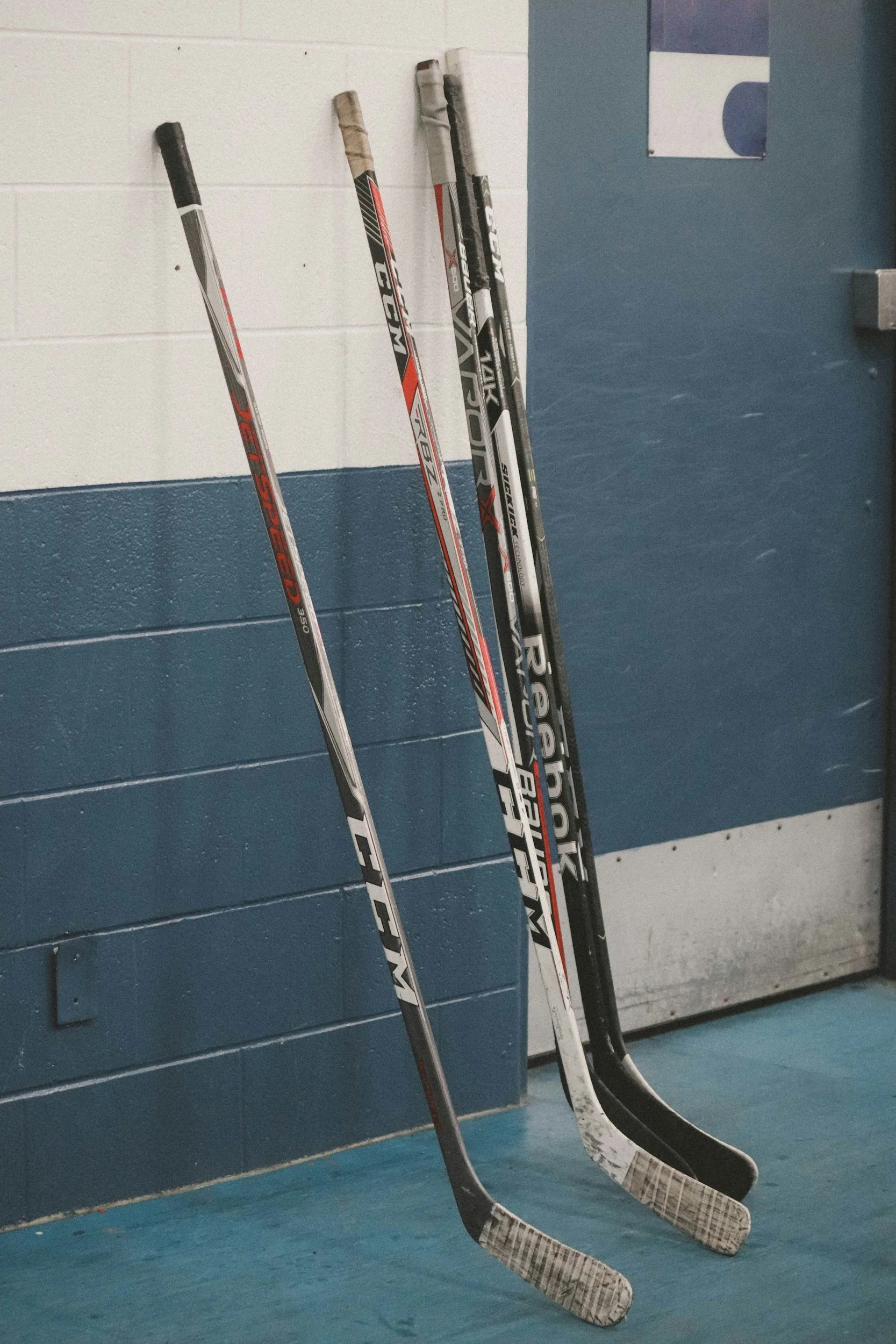 Three ice hockey sticks leaning against a wall in a locker room or ice rink, with a blue and white painted wall background.