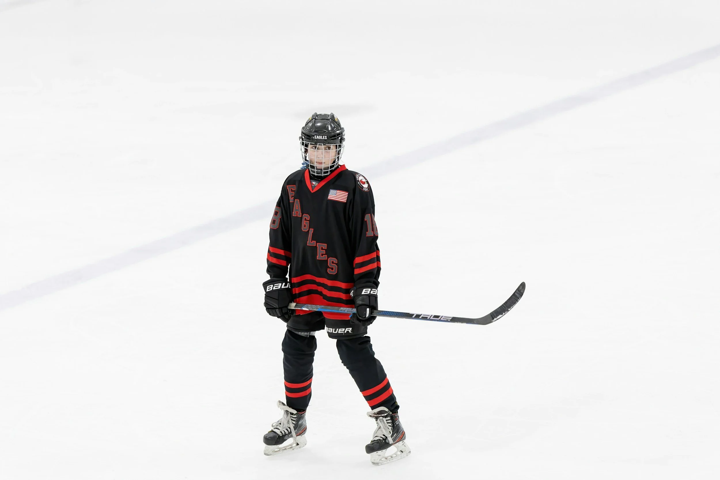 A young hockey player standing on the ice rink holding a hockey stick, wearing a black and red uniform with a helmet, gloves, and skates.