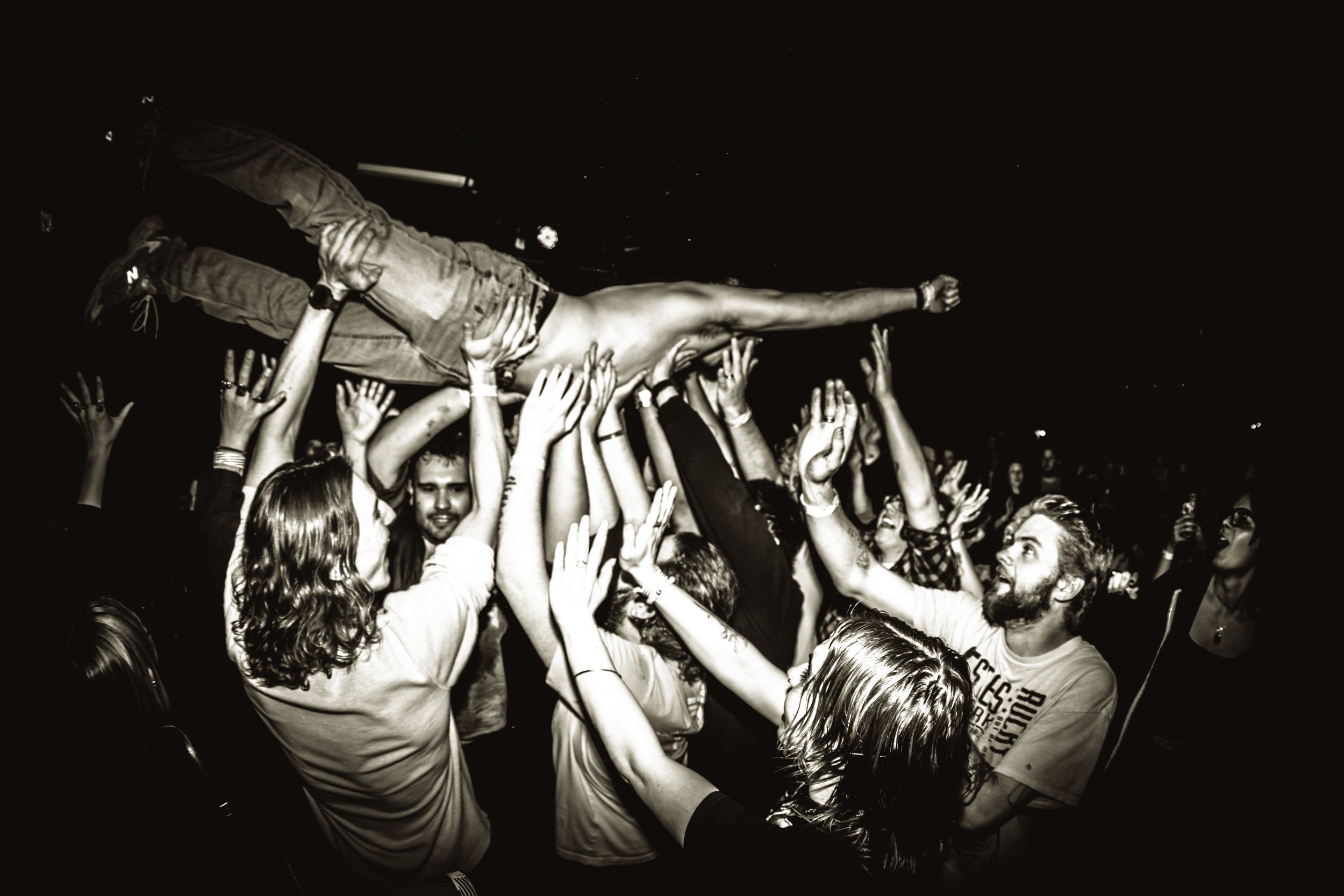 Black and white photo of a crowd at a concert or festival, people holding someone horizontally across the crowd, raising their hands, with a dark night sky in the background.