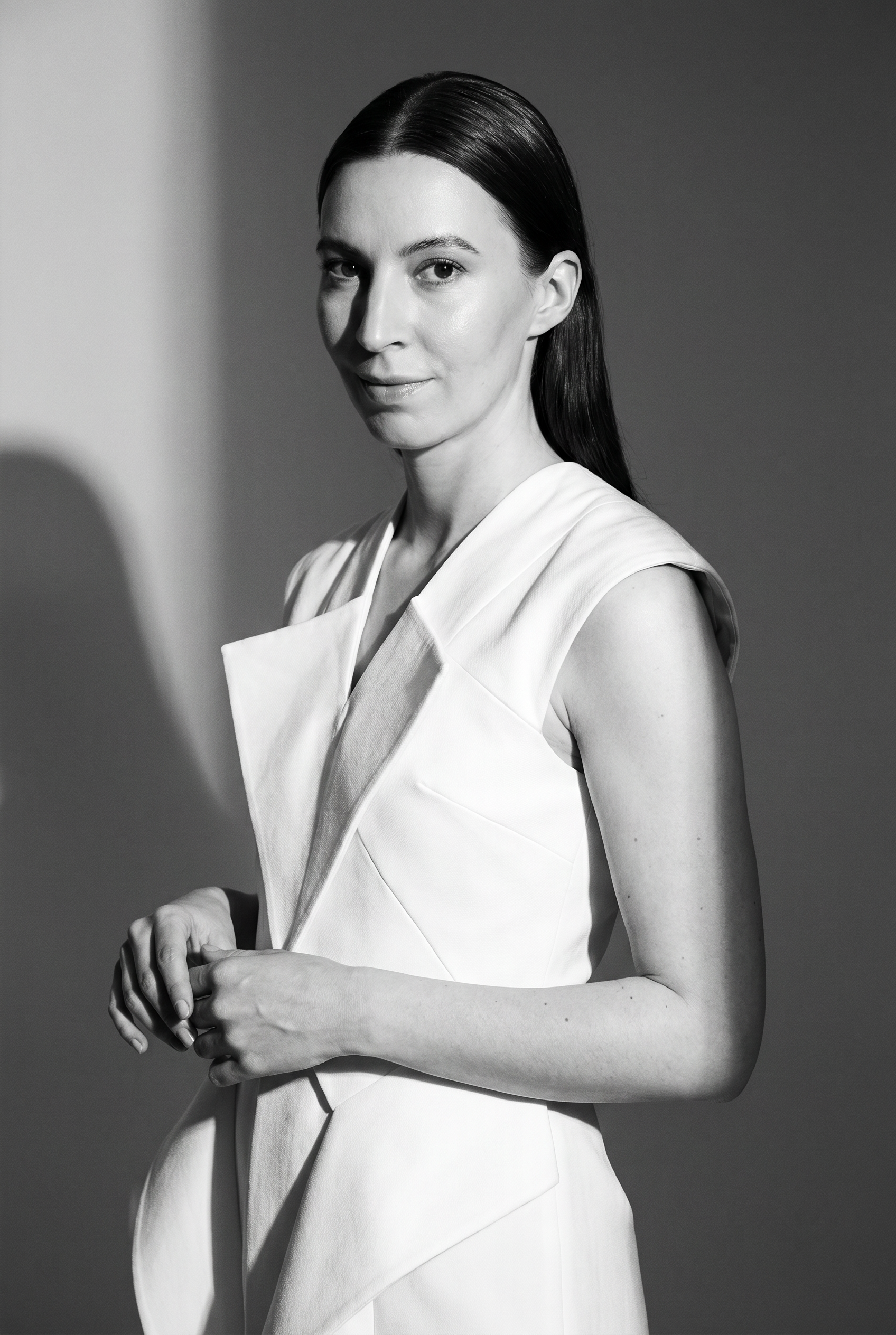 A woman with shoulder-length dark hair, wearing a sleeveless beige top and brown pants, sitting on a white bench against a wooden slat wall, writing in a notebook, with a framed abstract artwork and a bright, sunlit wall in the background.