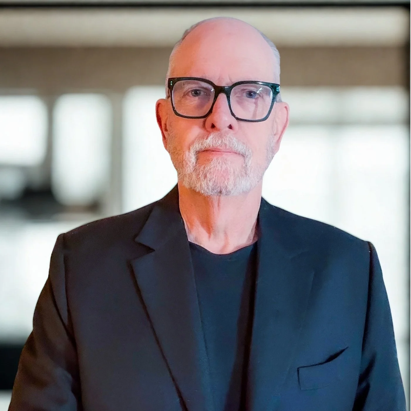A mature man with a beard and glasses wearing a black suit jacket, standing in front of a blurred background with natural light.