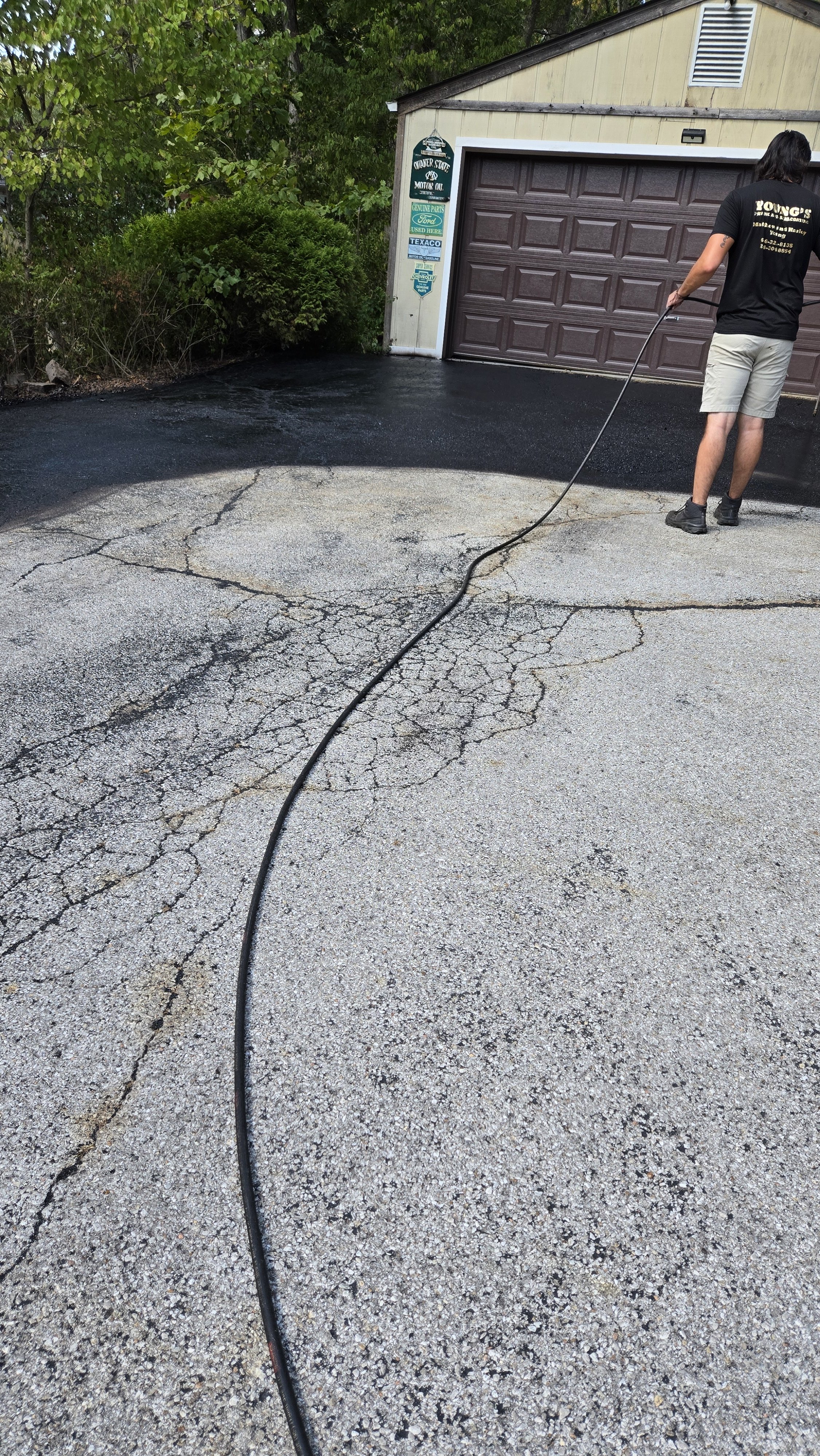 Person using a pressure washer to clean a cracked driveway in front of a garage, with greenery and a house in the background.