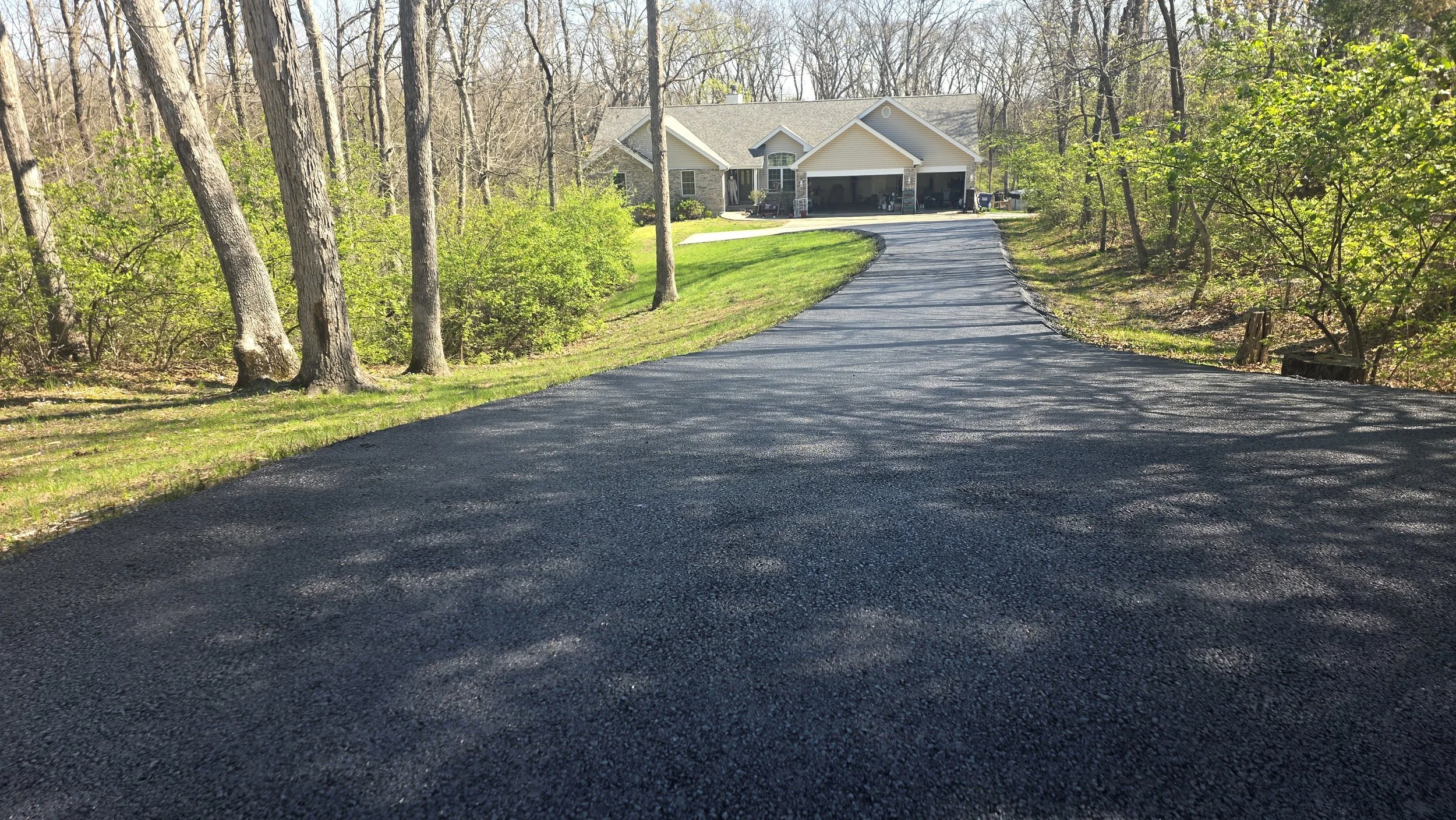 Newly paved black asphalt driveway leading to a house surrounded by trees and greenery on a sunny day.