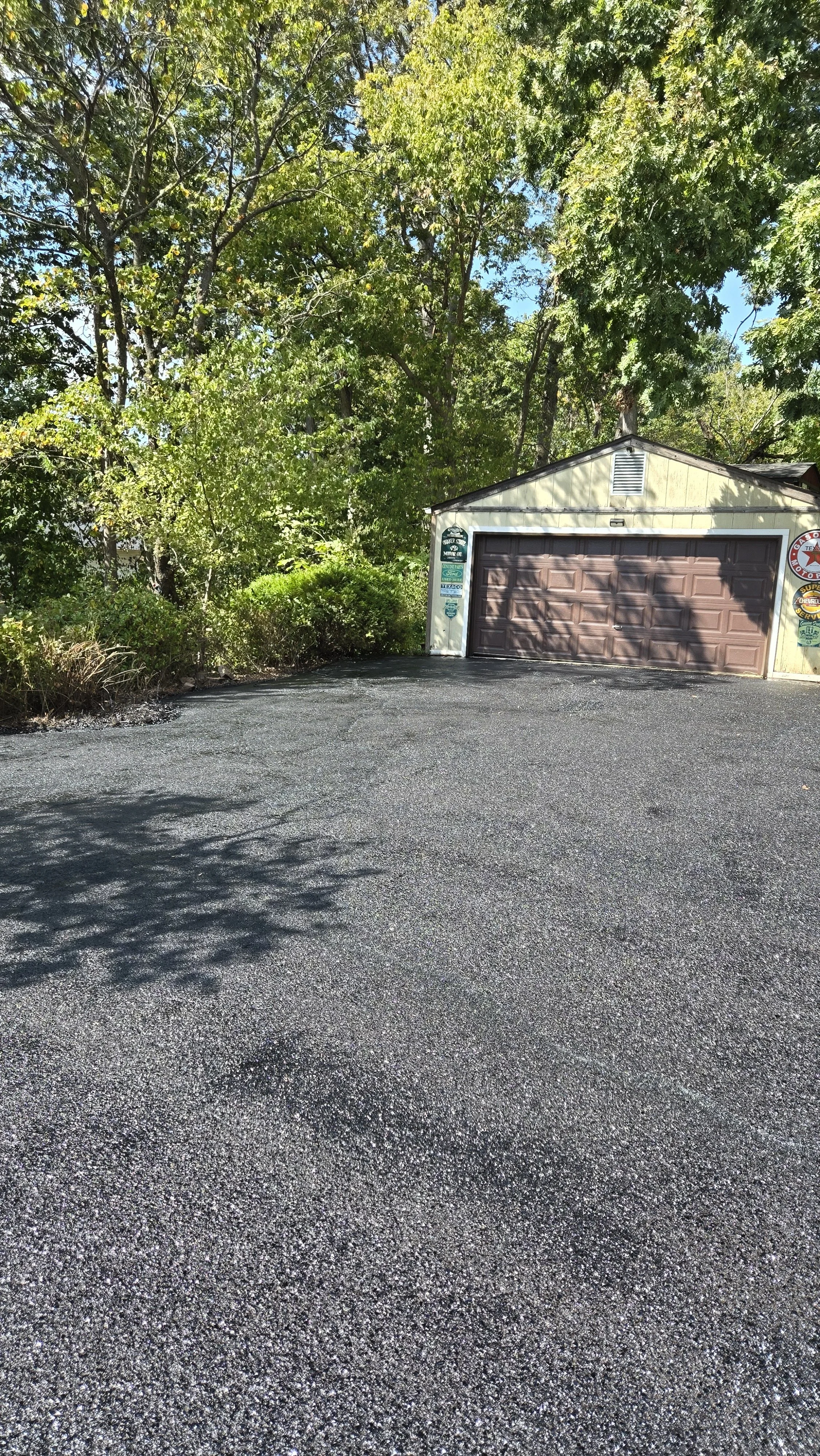 A gravel driveway leading to a small garage with a brown door, surrounded by trees and bushes with bright green leaves, andclear blue sky overhead.