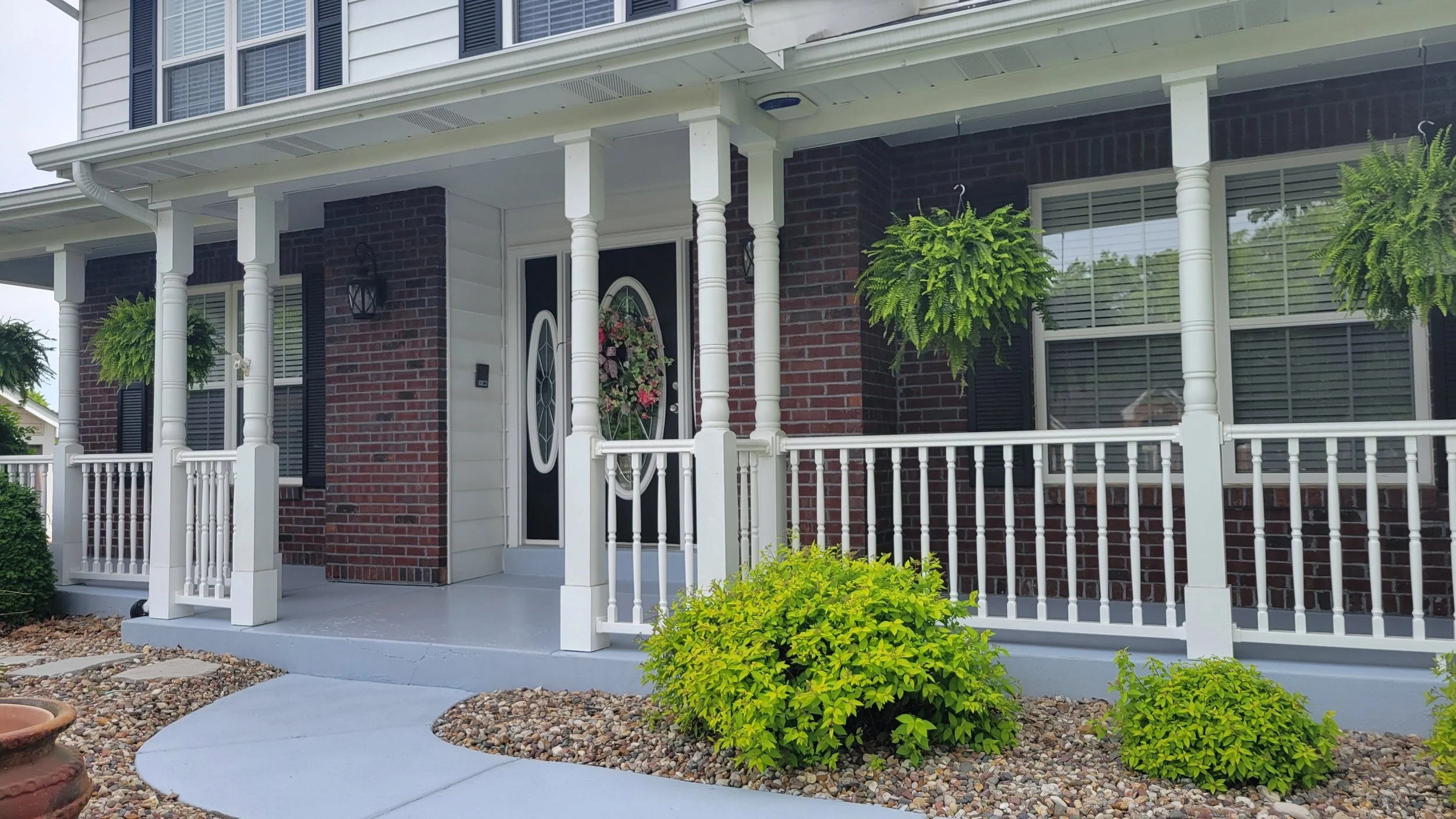 Front porch of a house with a black door decorated with a flower wreath, white railing, and hanging plants with green foliage, brick and white siding exterior