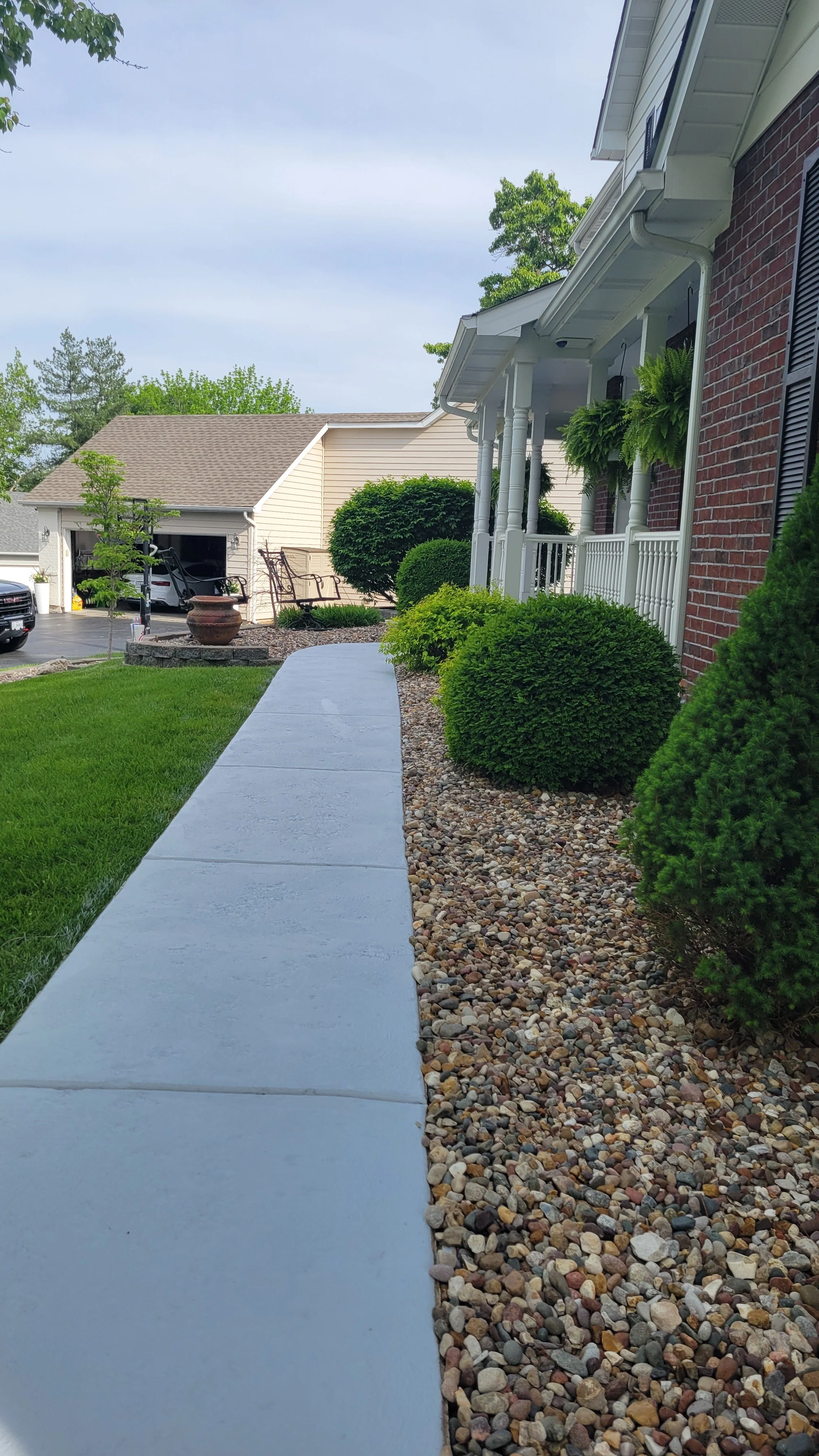 View of a front yard with a concrete walkway, landscaped with bushes, rocks, and a small tree, leading up to a porch of a brick house with hanging plants. In the background, there is a driveway with parked cars and a detached garage.