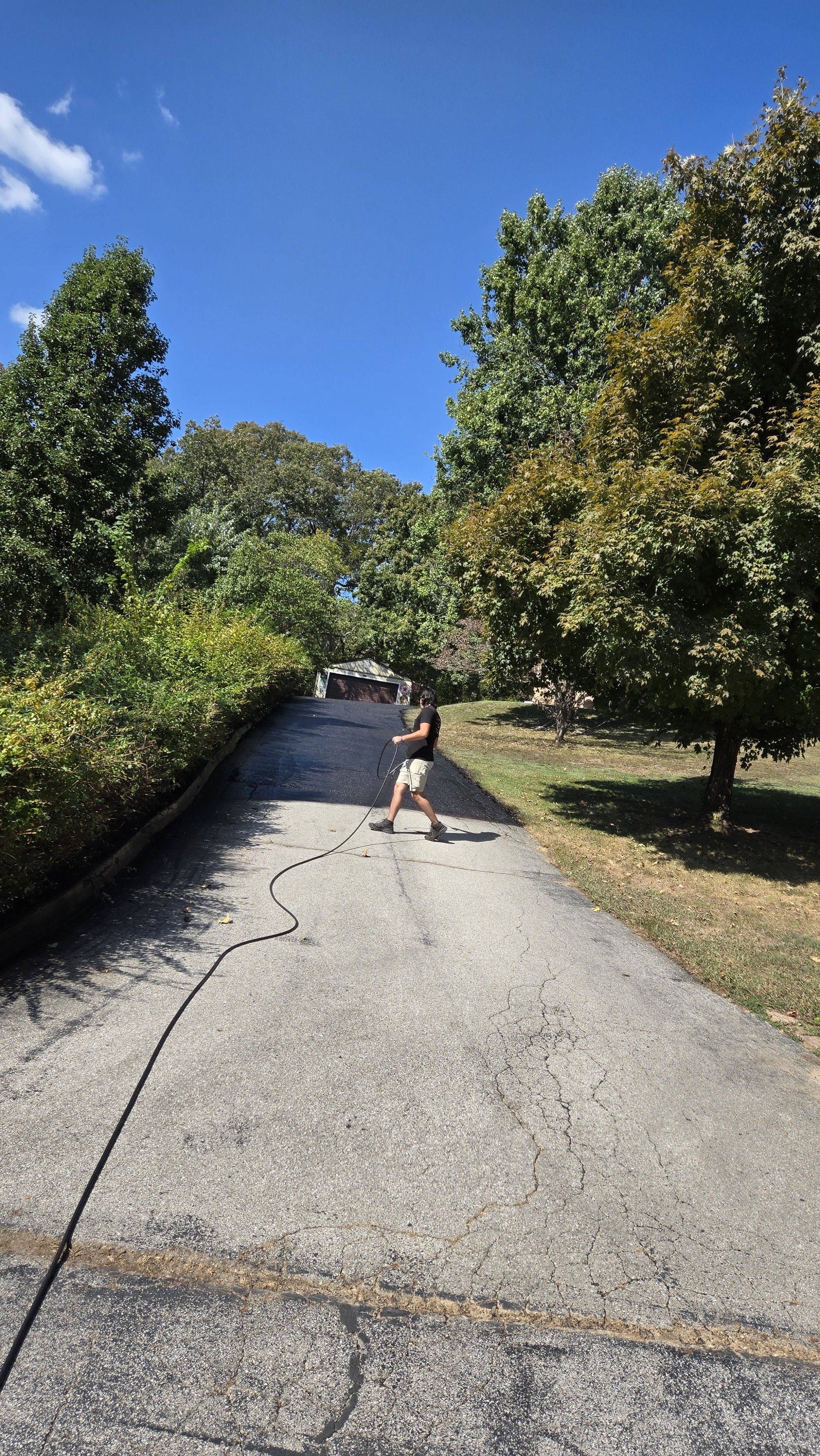 A person cleaning or pressure washing a driveway with a hose on a sunny day, with trees and a blue sky in the background.