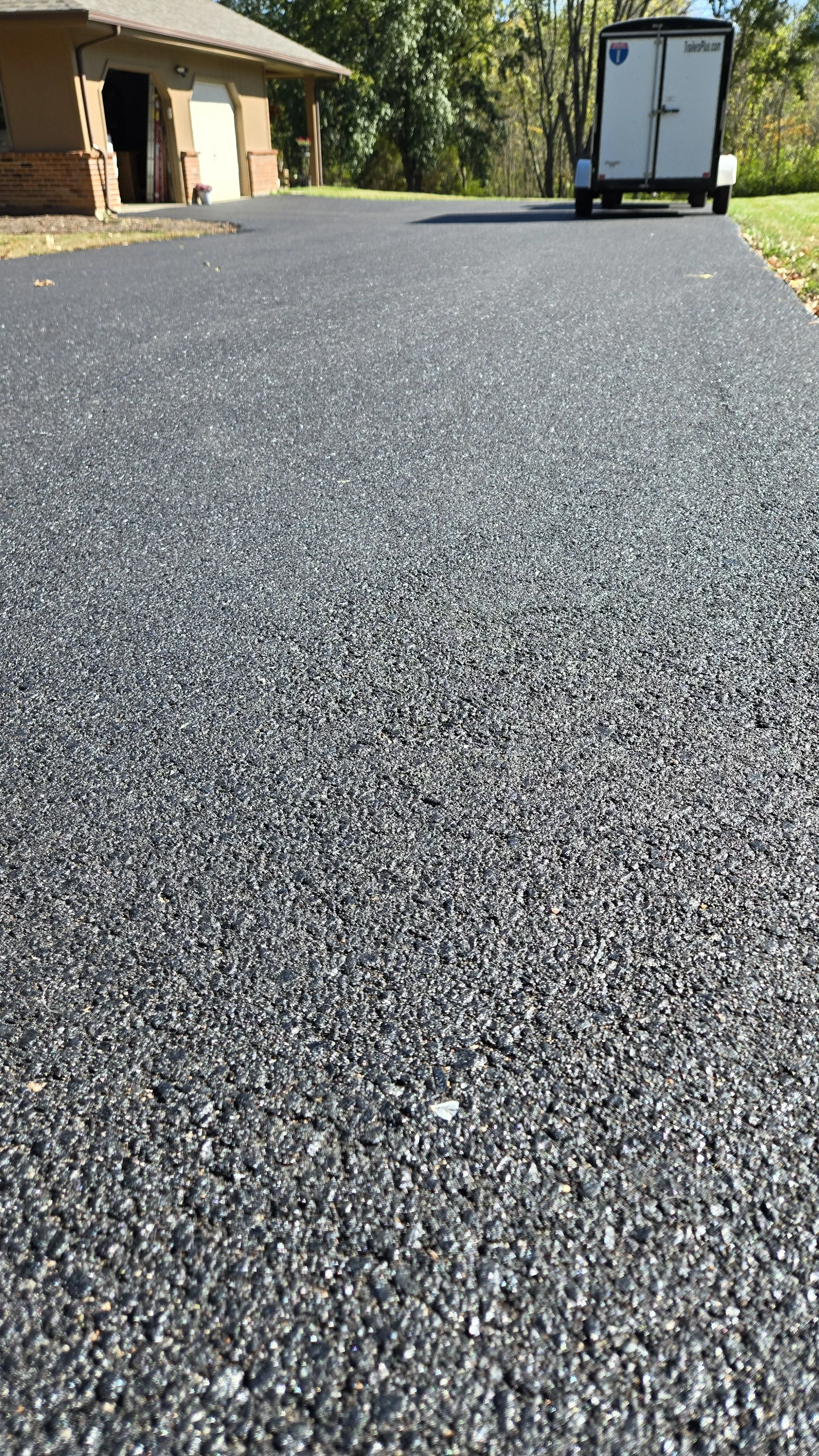 Close-up of a freshly paved asphalt driveway leading up to a house with a garage and a white utility trailer in the background, surrounded by trees and grass.
