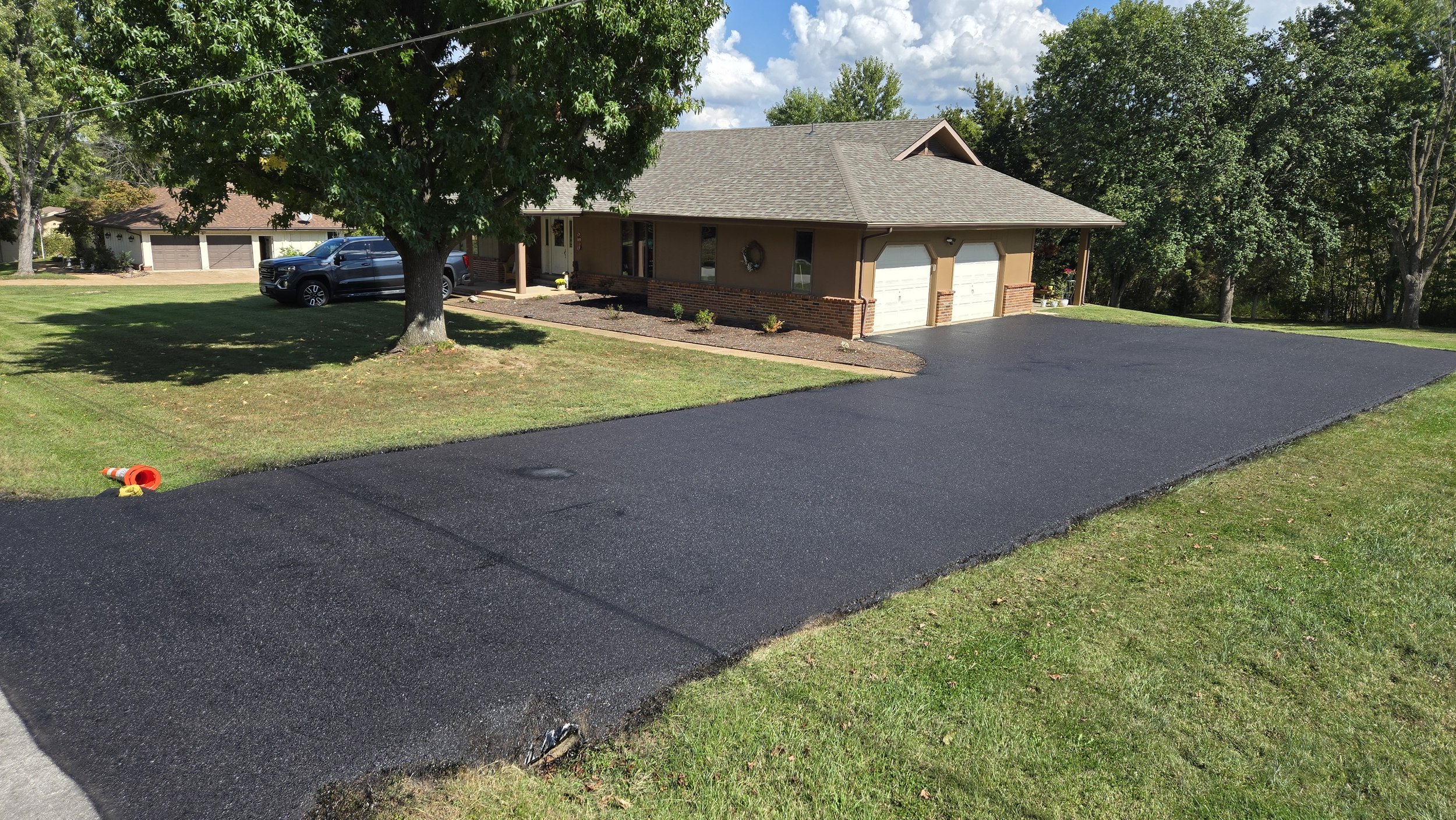 A newly paved black asphalt driveway in front of a beige house with two garage doors, a large tree, and a lawn with grass and small bushes, under a partly cloudy sky.
