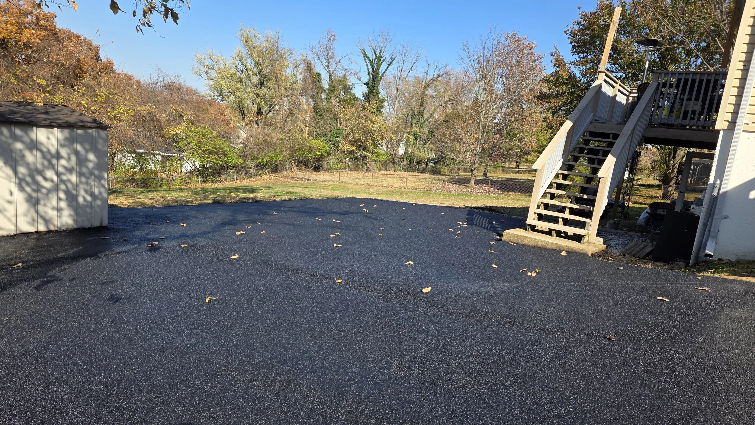 A backyard with a gravel driveway, a small storage shed, a wooden staircase leading to a deck, and trees with fall foliage in the background under a clear blue sky.