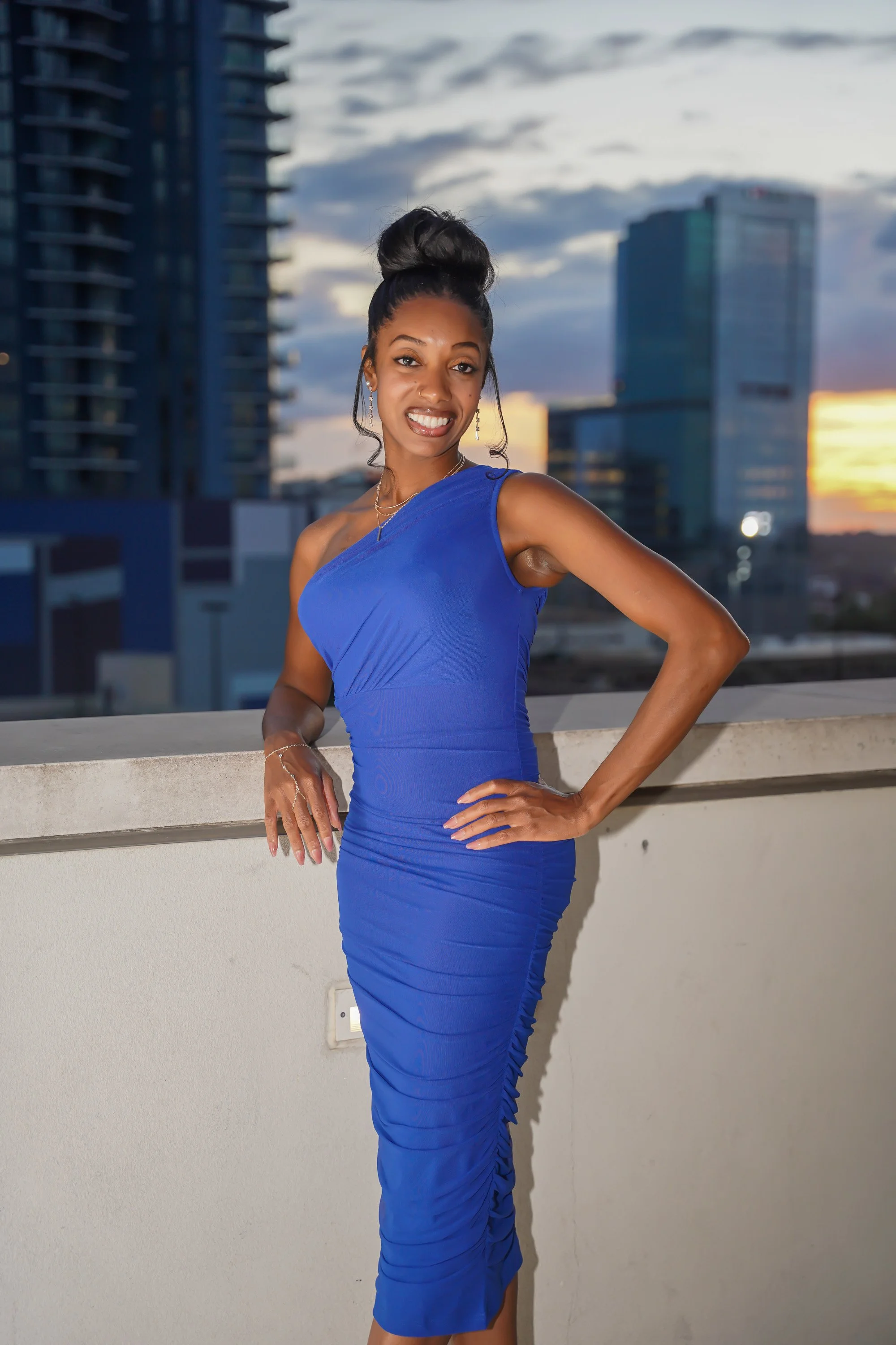 A woman in a royal blue dress standing on a balcony with city buildings and a sunset sky in the background.