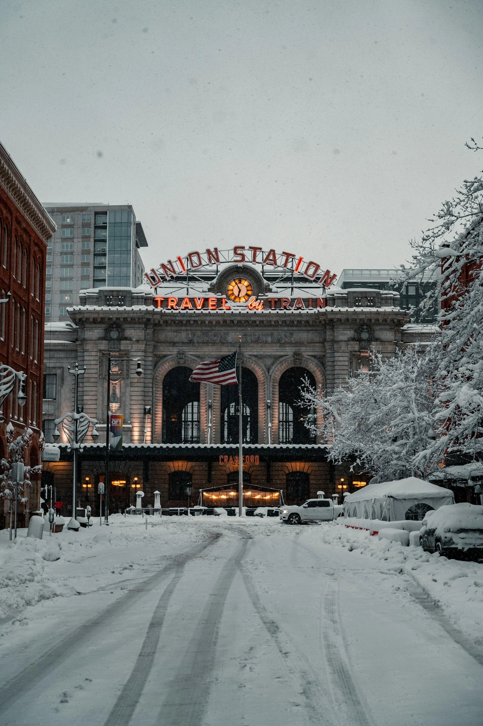The exterior of a train station, Union Station, on a snowy day, with a large illuminated sign reading 'Union Station Travel by Train', a flag in front, and modern buildings in the background.