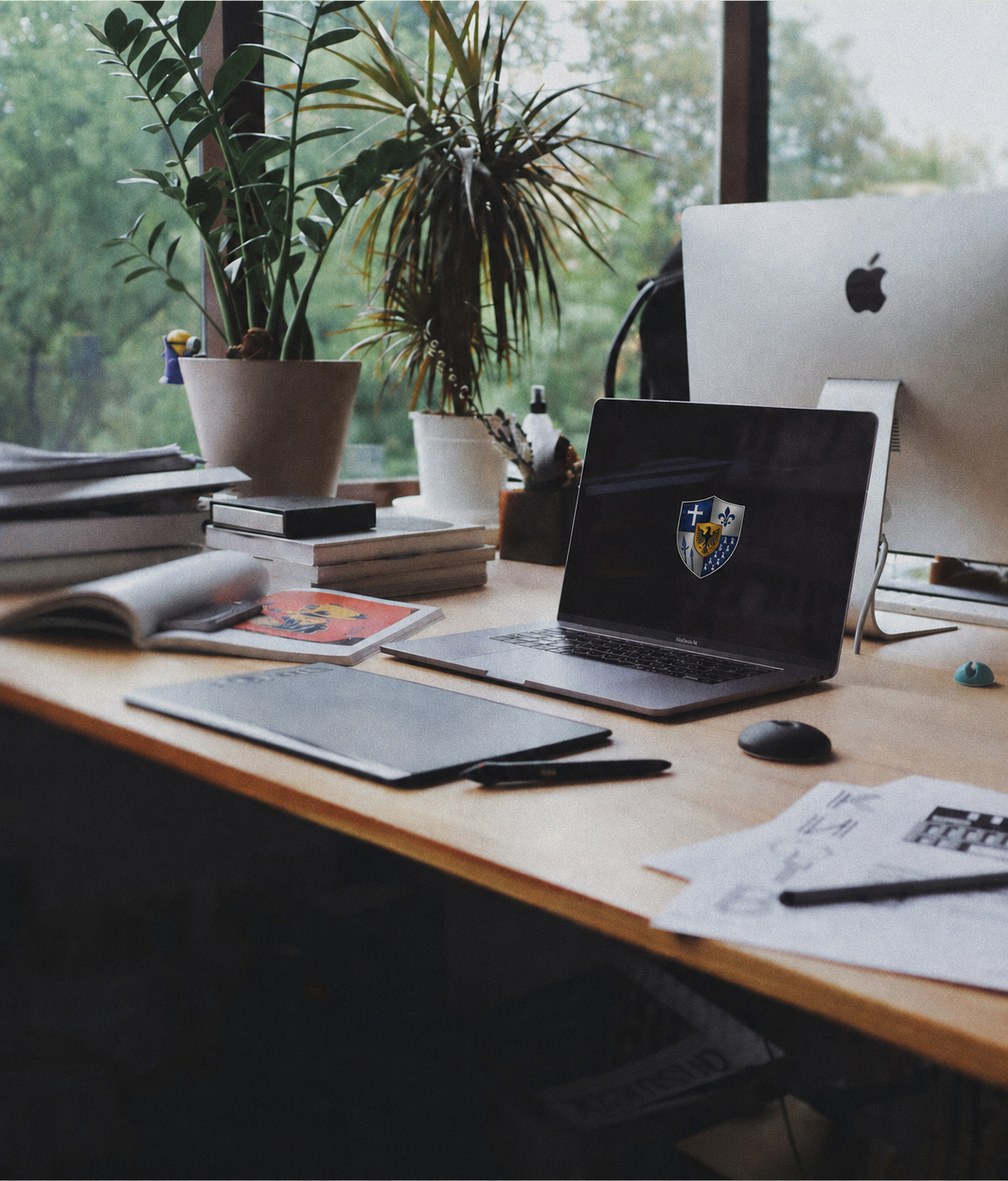 A cluttered workspace with two potted plants, a laptop displaying the HCTS logo, an Apple desktop computer, notebooks, papers, pens, and a computer mouse on a wooden desk by a window with greenery outside.