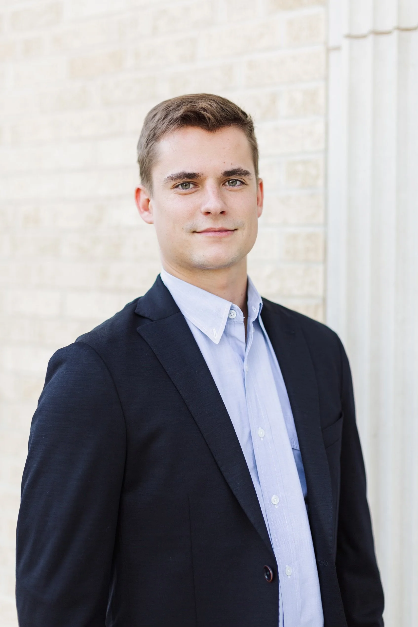 A young man in business attire standing outdoors against a light-colored brick wall.