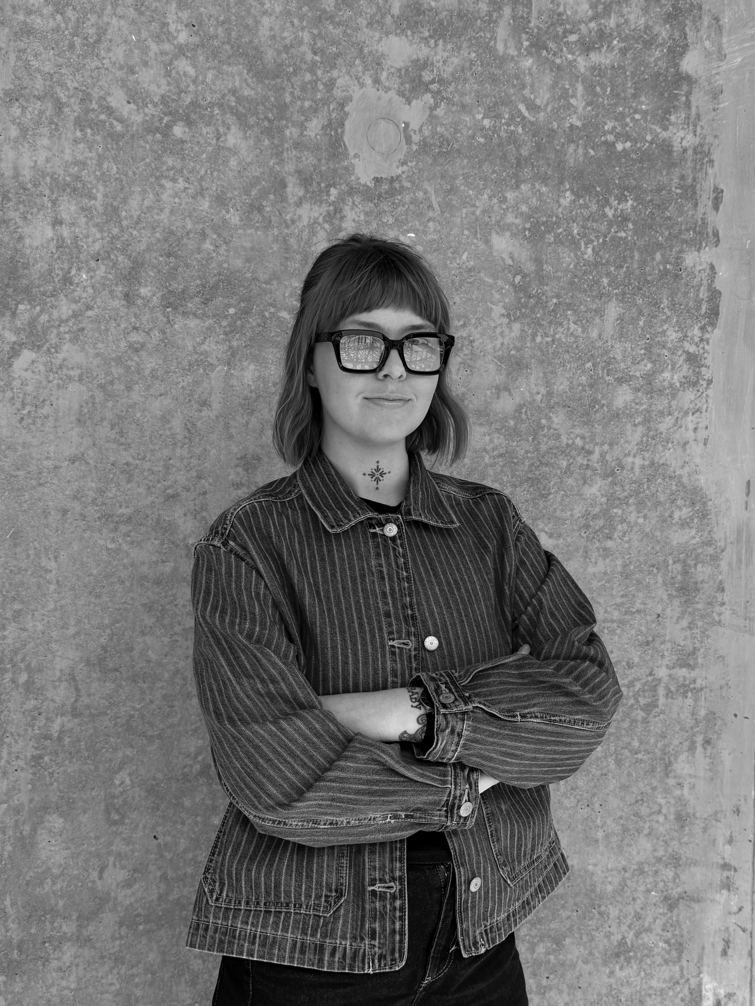 A woman with shoulder-length hair, wearing large glasses, and a denim jacket, stands against a textured wall with her arms crossed.