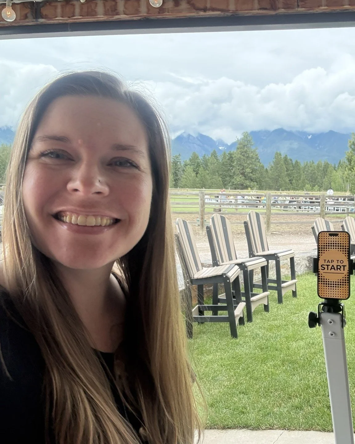 A woman smiling inside a building with a view of mountains, trees, and chairs outside, and a smartphone with a 'Tap to Start' sign on a stand in the foreground.