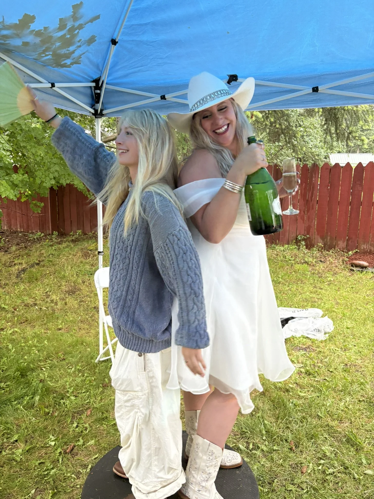 Two women standing back-to-back outdoors on a round platform under a blue canopy, holding drinks, one in a cowboy hat and the other in a sweater, smiling and enjoying a festive moment.