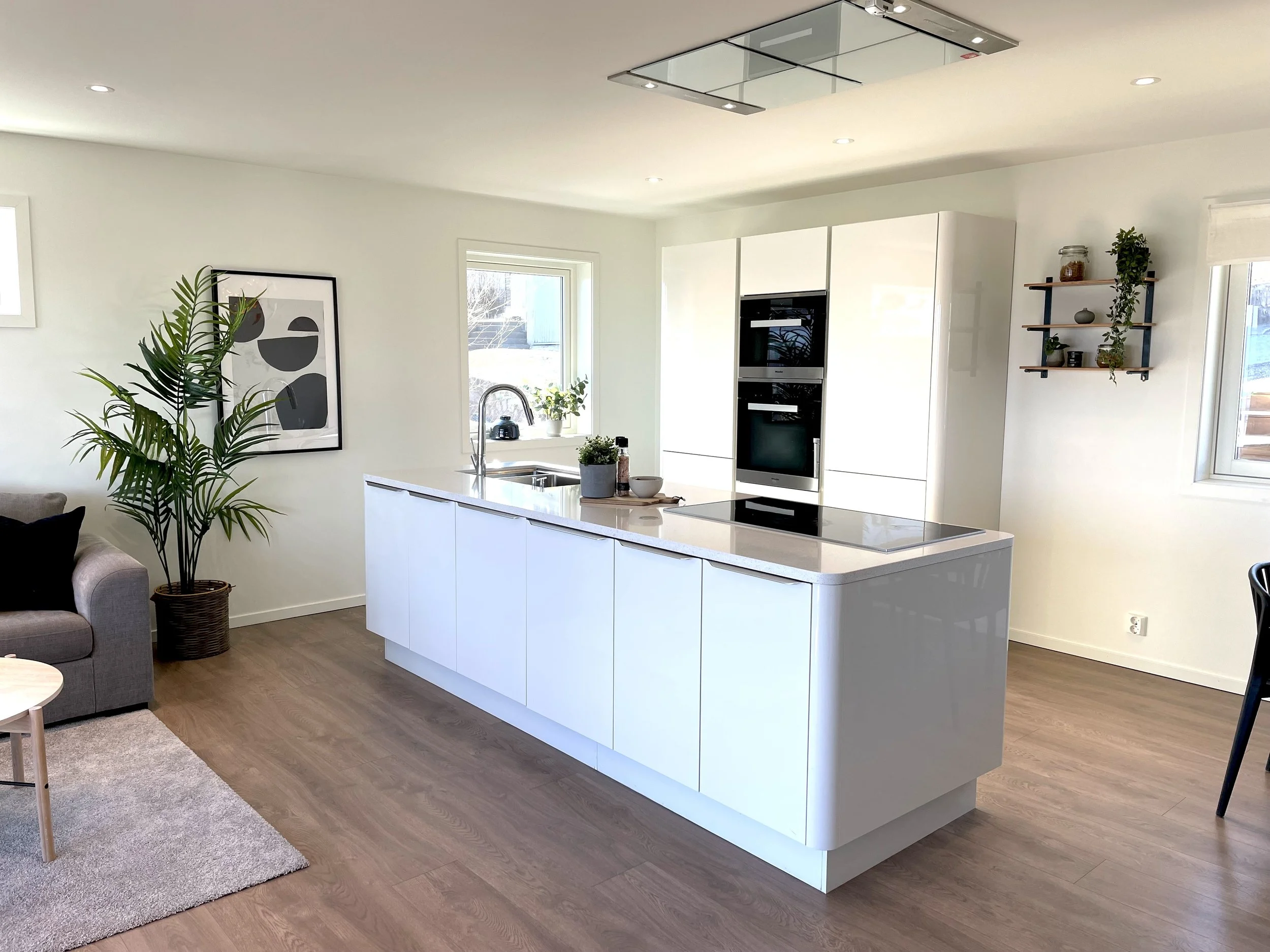 Interior view of a modern kitchen with white cabinetry, a kitchen island, a built-in oven, a potted plant, and a large window letting in natural light.