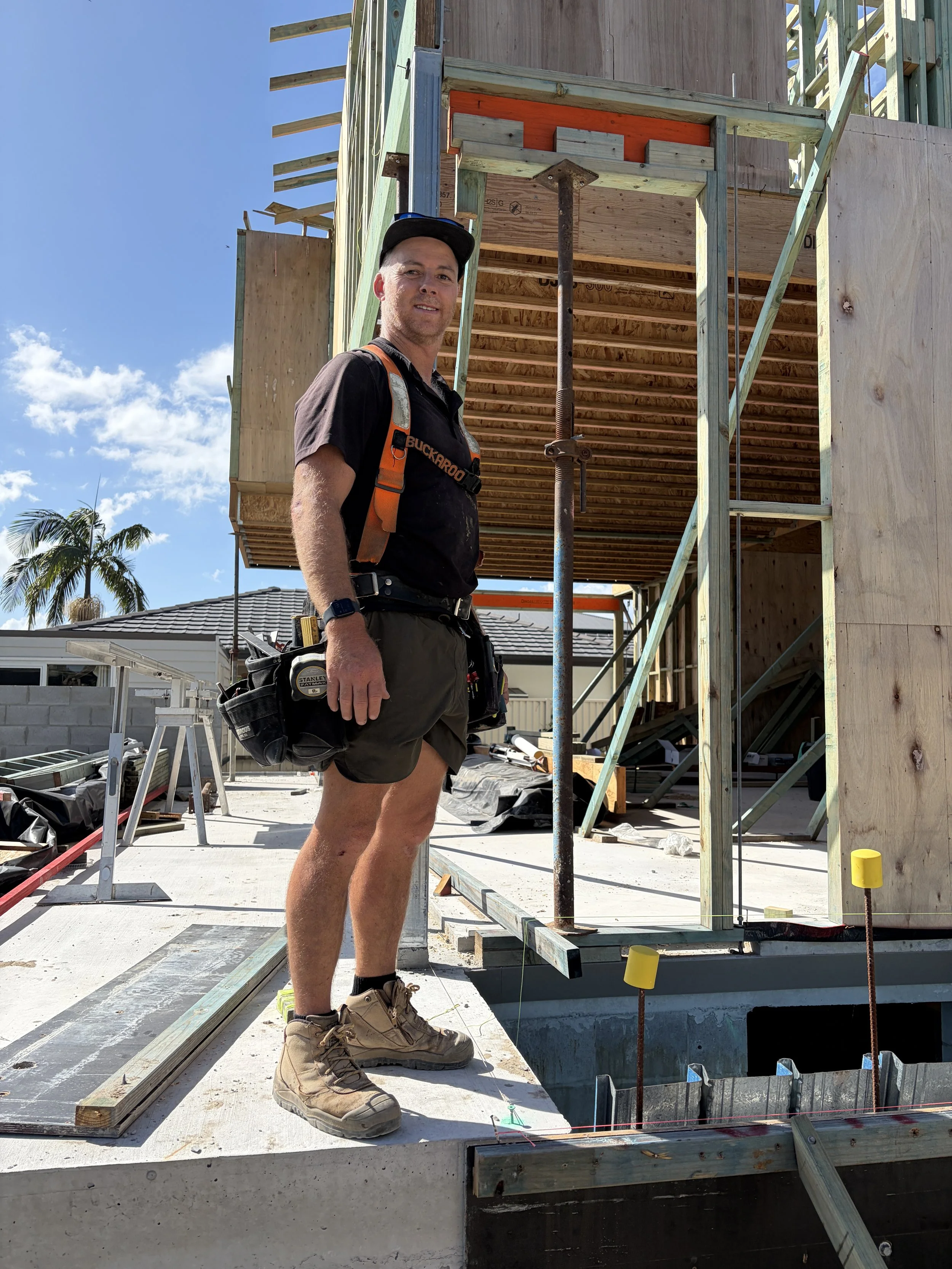 A construction worker standing on a concrete foundation at a building site, smiling, with a partly built wooden structure behind him under a clear blue sky.