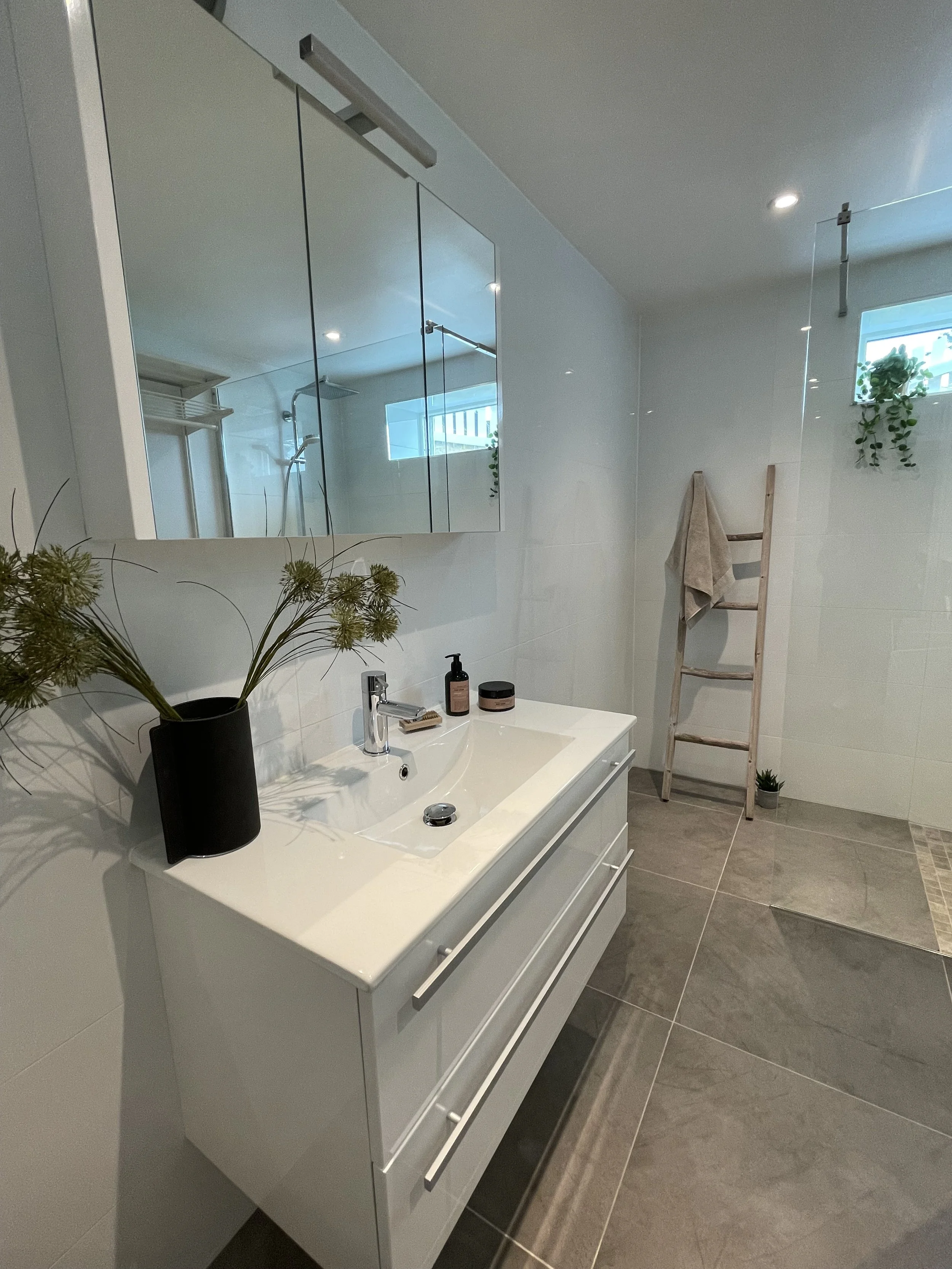 Modern bathroom with white vanity, large mirror, potted plant, and ladder with towel, with a shower visible in the mirror reflection and small window with plants.