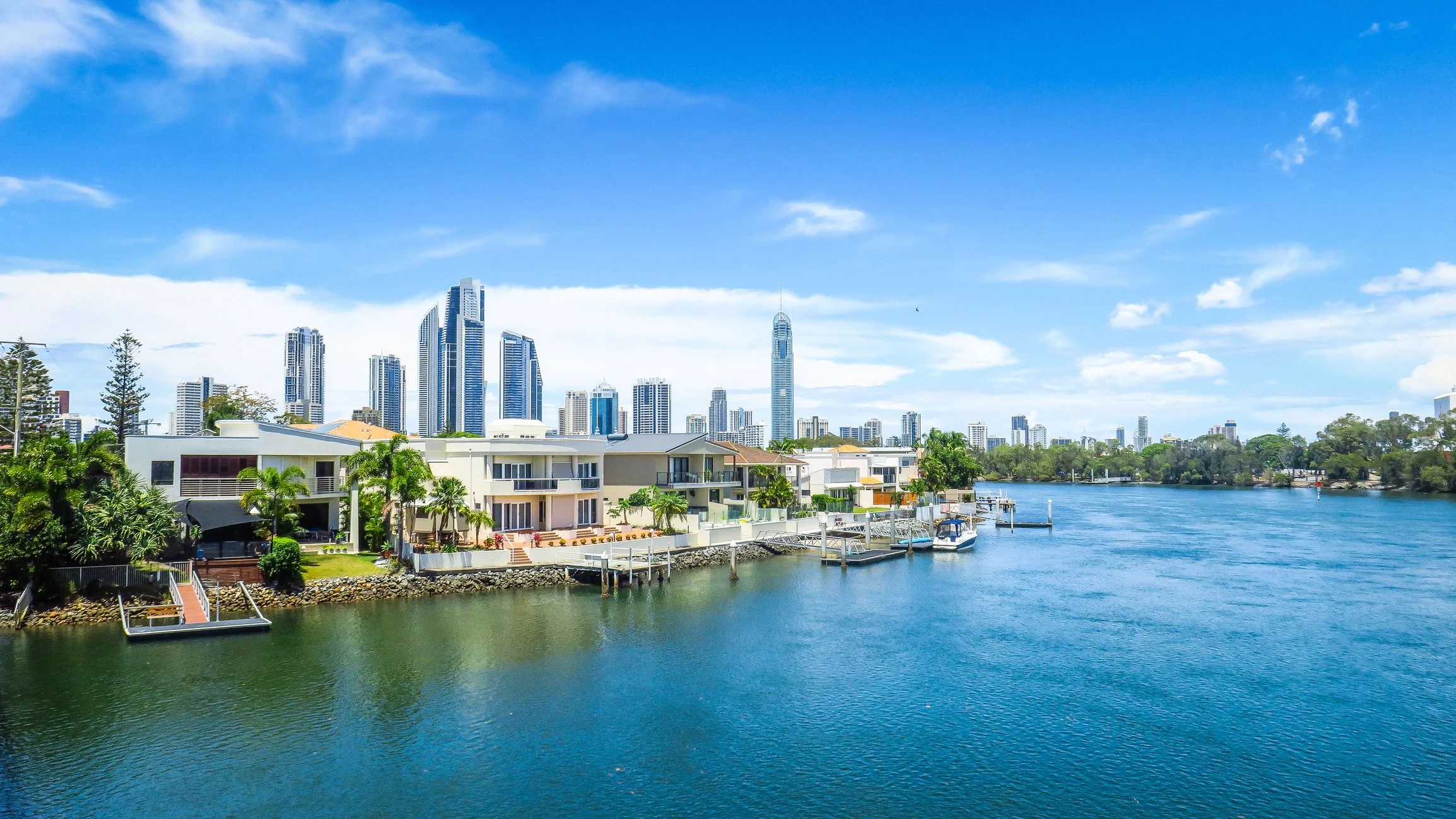 City skyline with tall modern skyscrapers across a river, with waterfront houses and boats, under a partly cloudy blue sky.