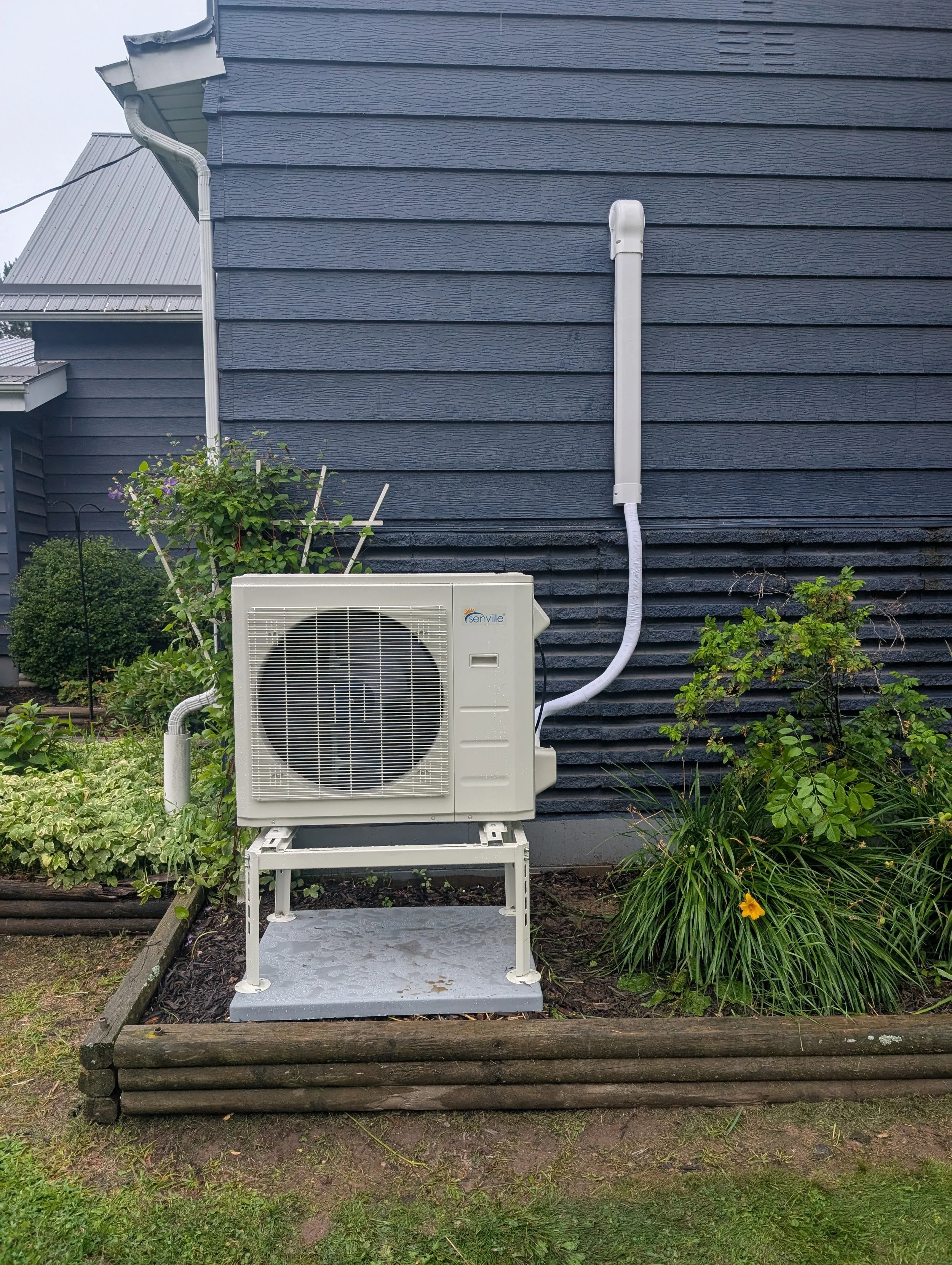 The image shows an outdoor scene with a dark blue house wall with horizontal siding, an air conditioning unit on a raised platform, white pipes, and surrounding green plants with some yellow flowers. There is a garden bed bordered by wooden logs at the base of the house.