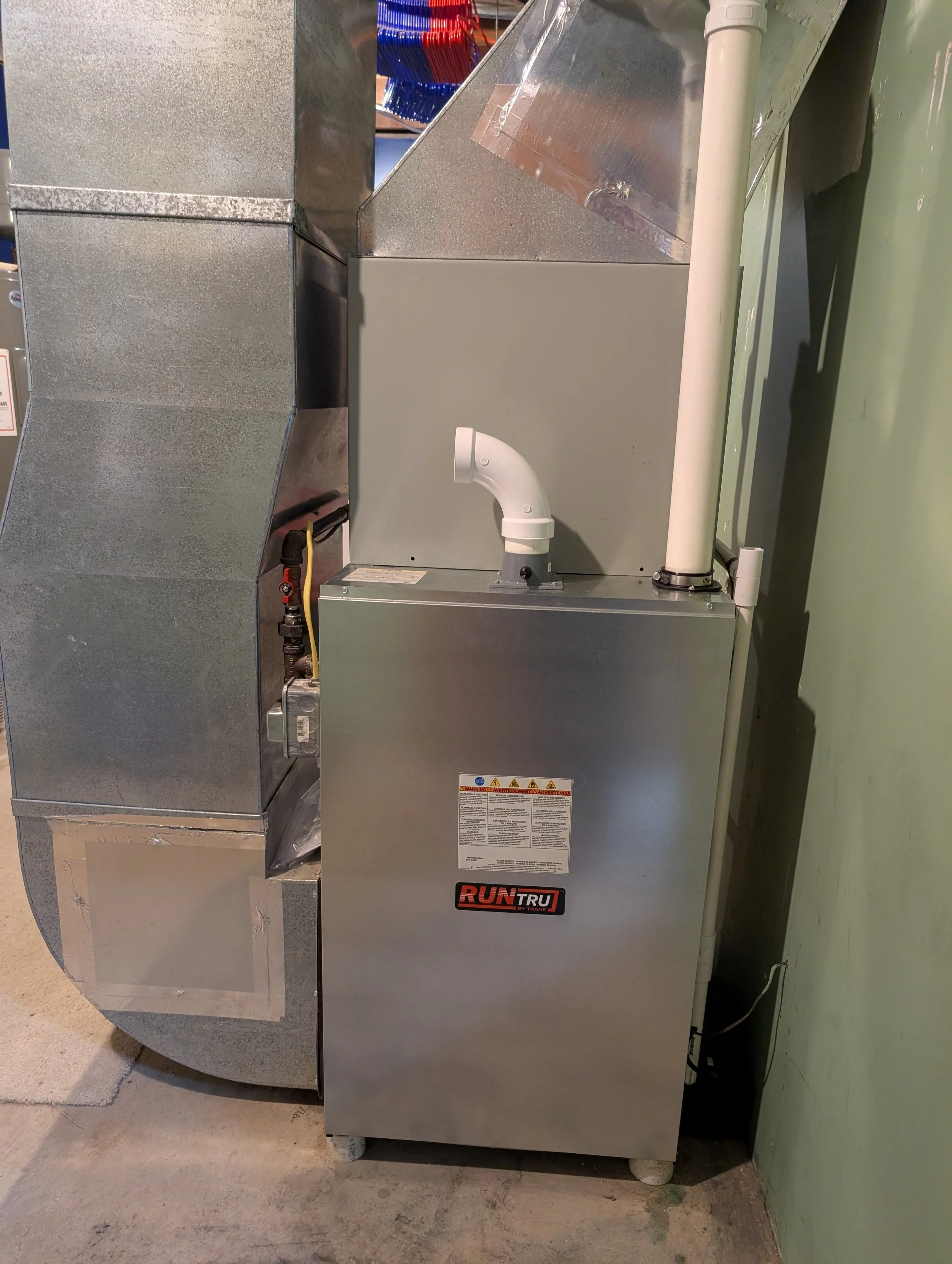 Metal furnace or heating unit labeled 'Run Tru' with white PVC pipe and a warning label, positioned against a green wall in a concrete room.