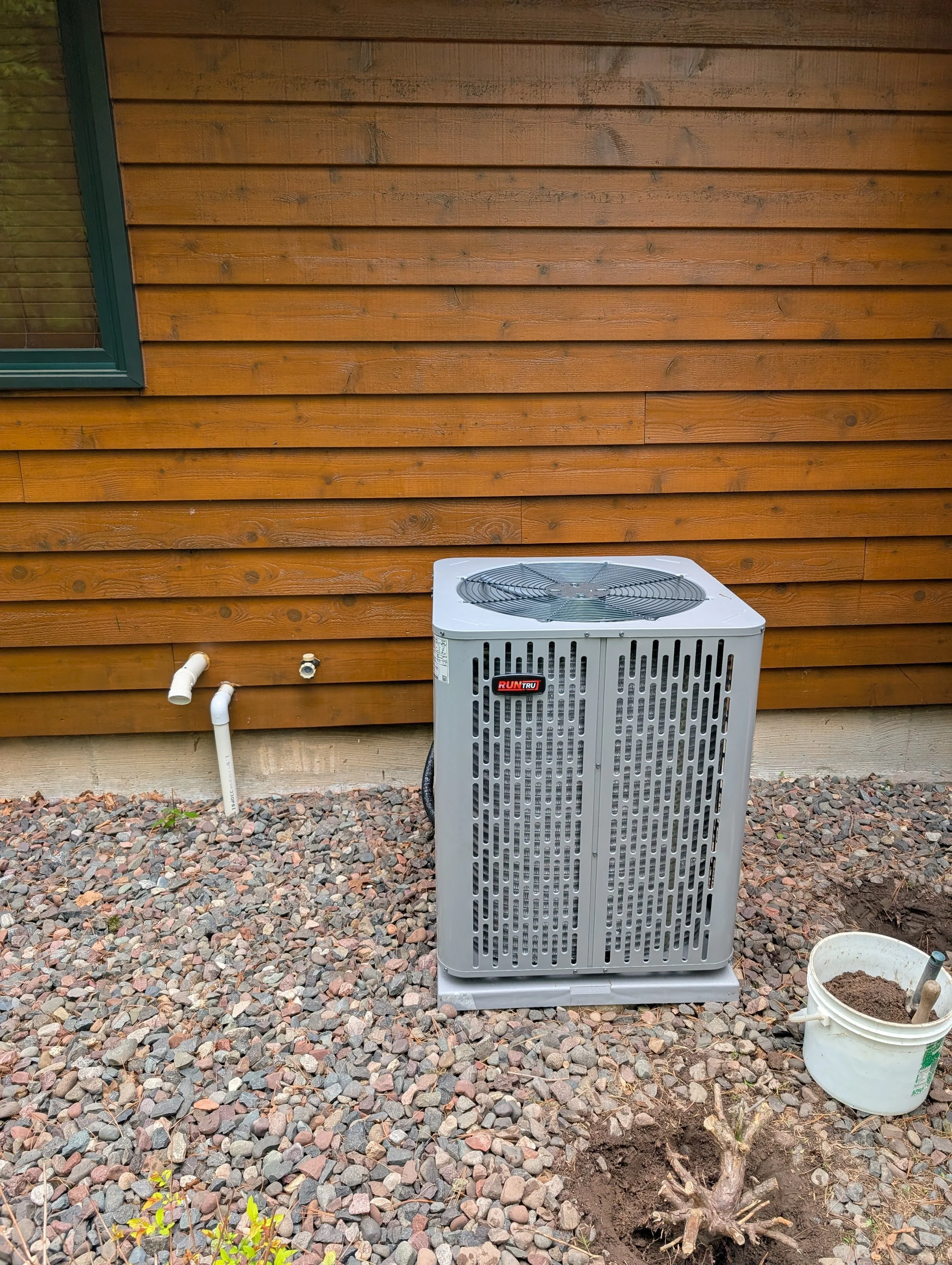 An outdoor scene with brown wooden wall siding, a window with blinds, a white HVAC unit, a white pipe connection, and a bucket with soil and a small plant on rocky ground.